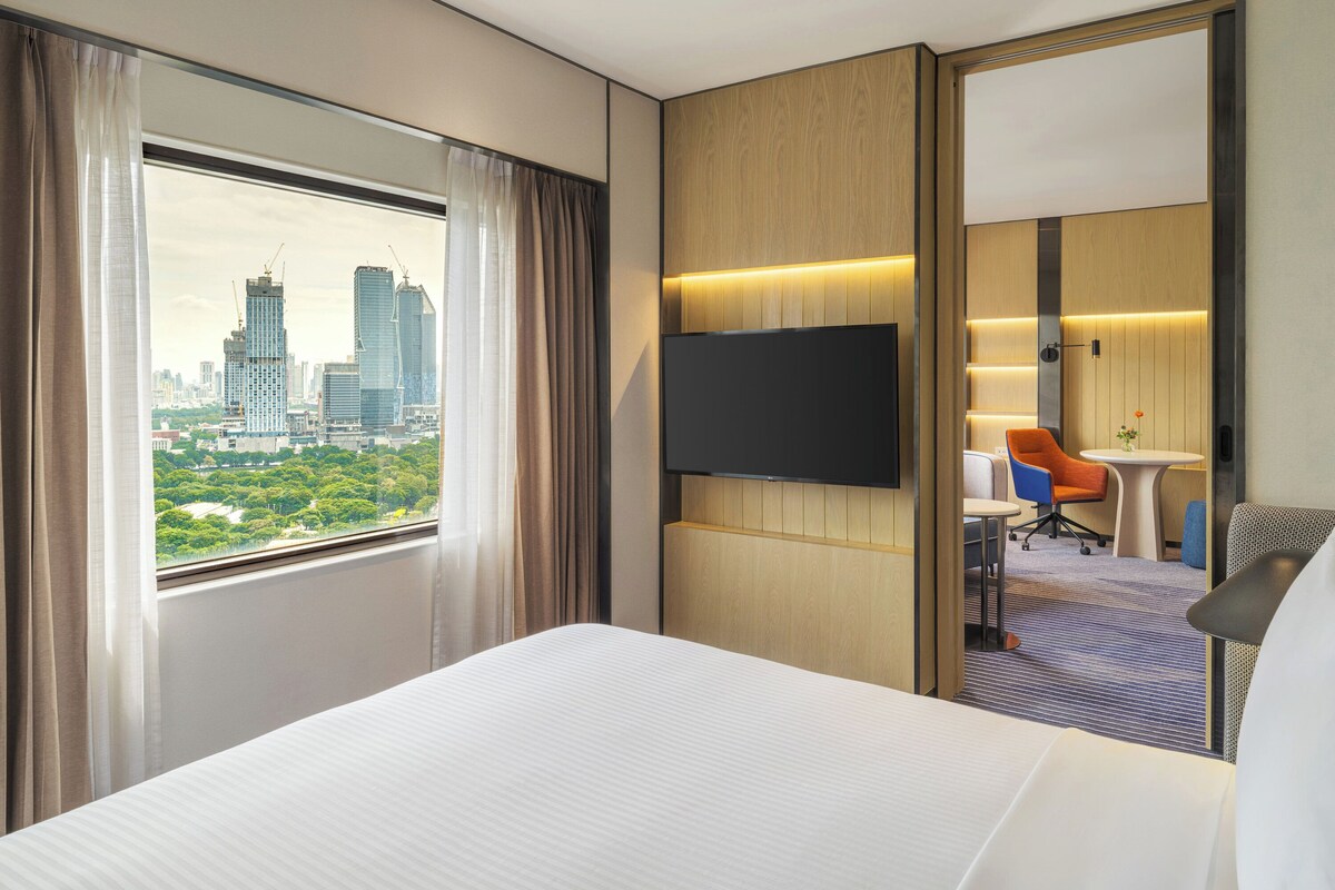 Habitación de hotel con una cama grande, un televisor en la pared, un escritorio con una silla y vistas a la ciudad desde la ventana.