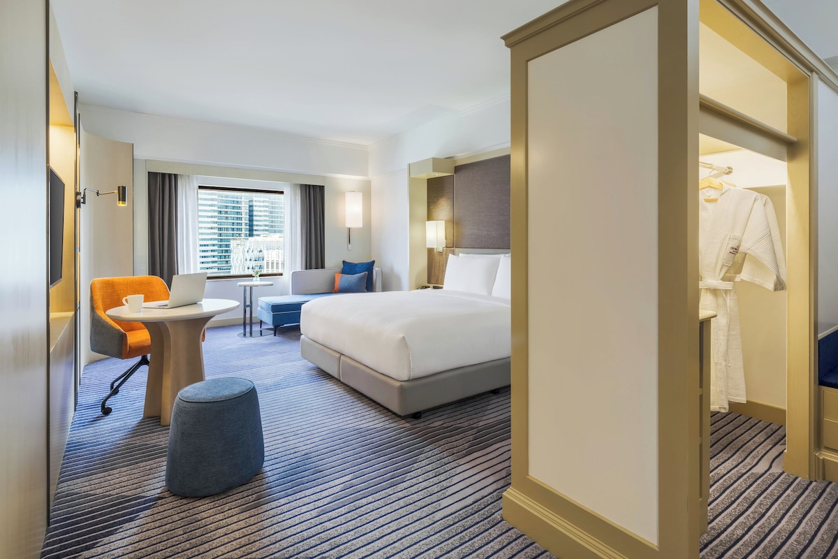 Habitación de hotel con cama, escritorio, silla, una mesita y ventana con vista a edificios.