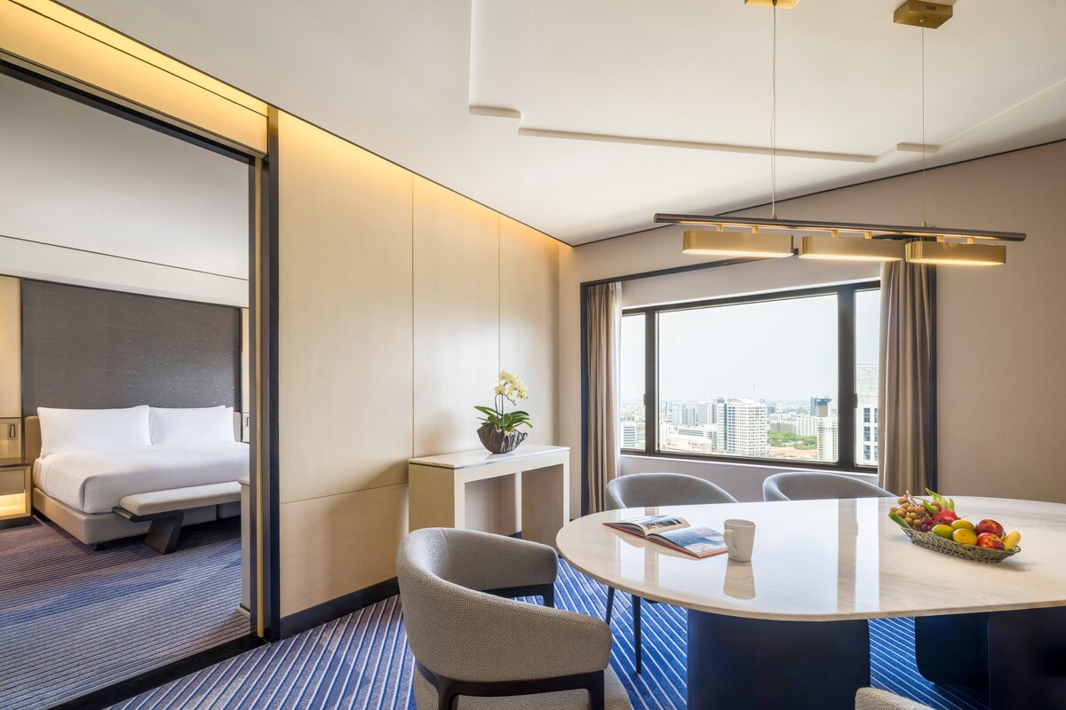 Habitación de hotel con cama, escritorio, silla, mesa con frutero y vista de la ciudad a través de amplias ventanas.