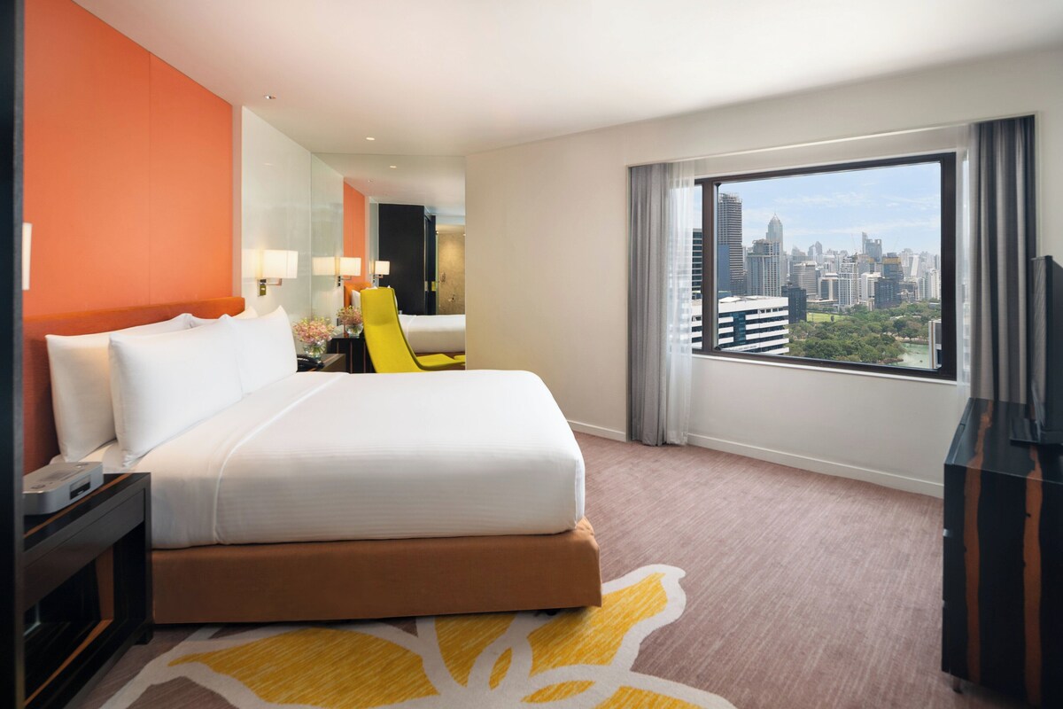 Habitación de hotel con cama, escritorio, silla, televisión y un amplio ventanal con vistas a la ciudad.