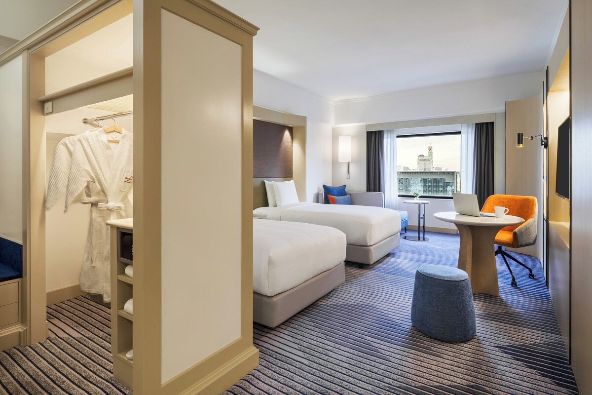 Habitación de hotel con cama, dos mesitas de noche, una silla, un escritorio con una computadora portátil y vistas a la ciudad.