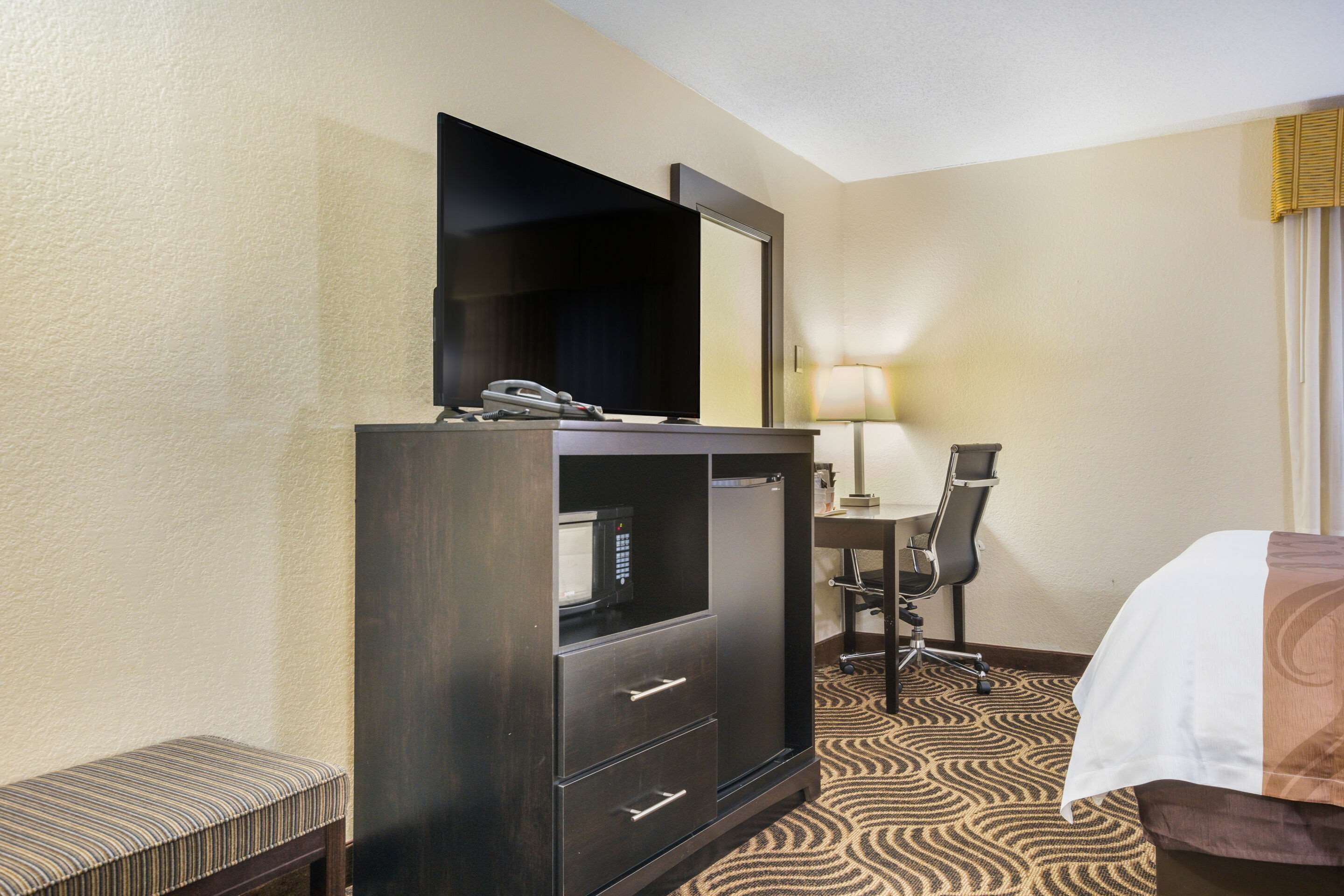 Een hotelkamer met een donker houten dressoir, een bureau met stoel, een bed met een wit-bruine sprei en een kamerbreed tapijt met dessin.