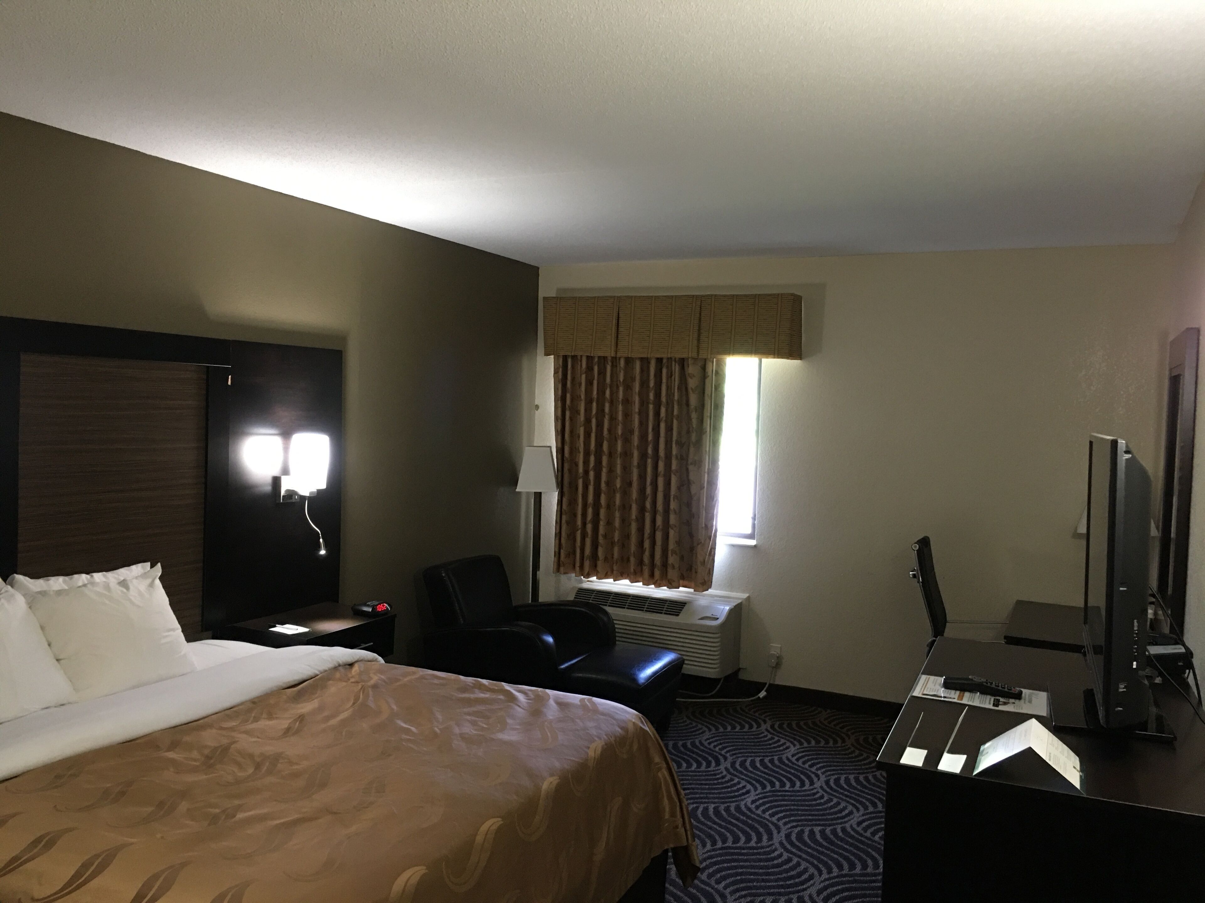 A hotel room with a bed, a desk with a computer, a chair, a window with curtains, and a wall-mounted TV.