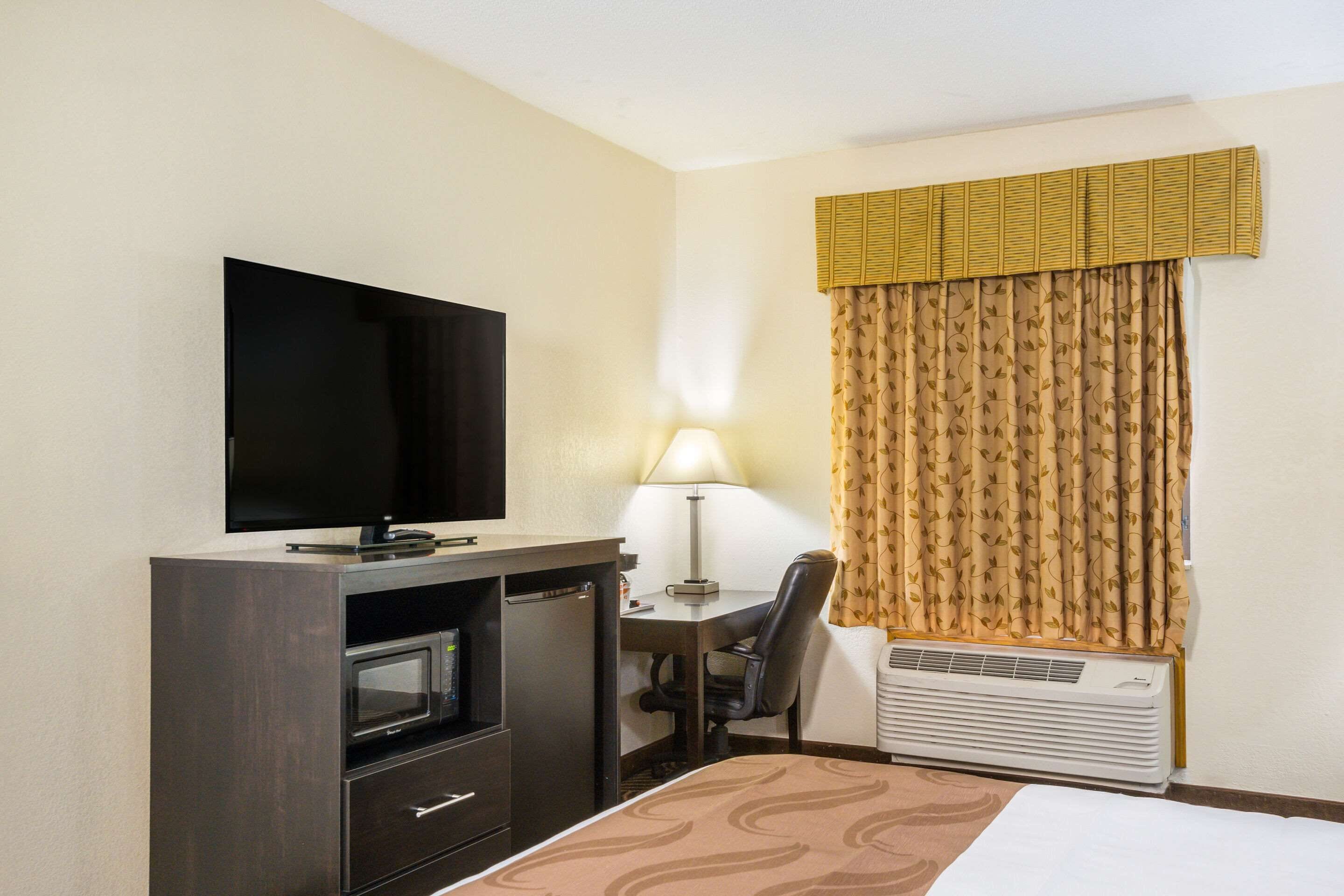 A hotel room with a flat-screen TV, a desk with a chair, a lamp, a bed with a patterned bedspread, a minibar, and a window with curtains.