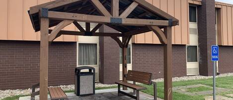 Un área de estar exterior cubierta con bancos y un contenedor de basura, contigua a un edificio con un letrero de estacionamiento.