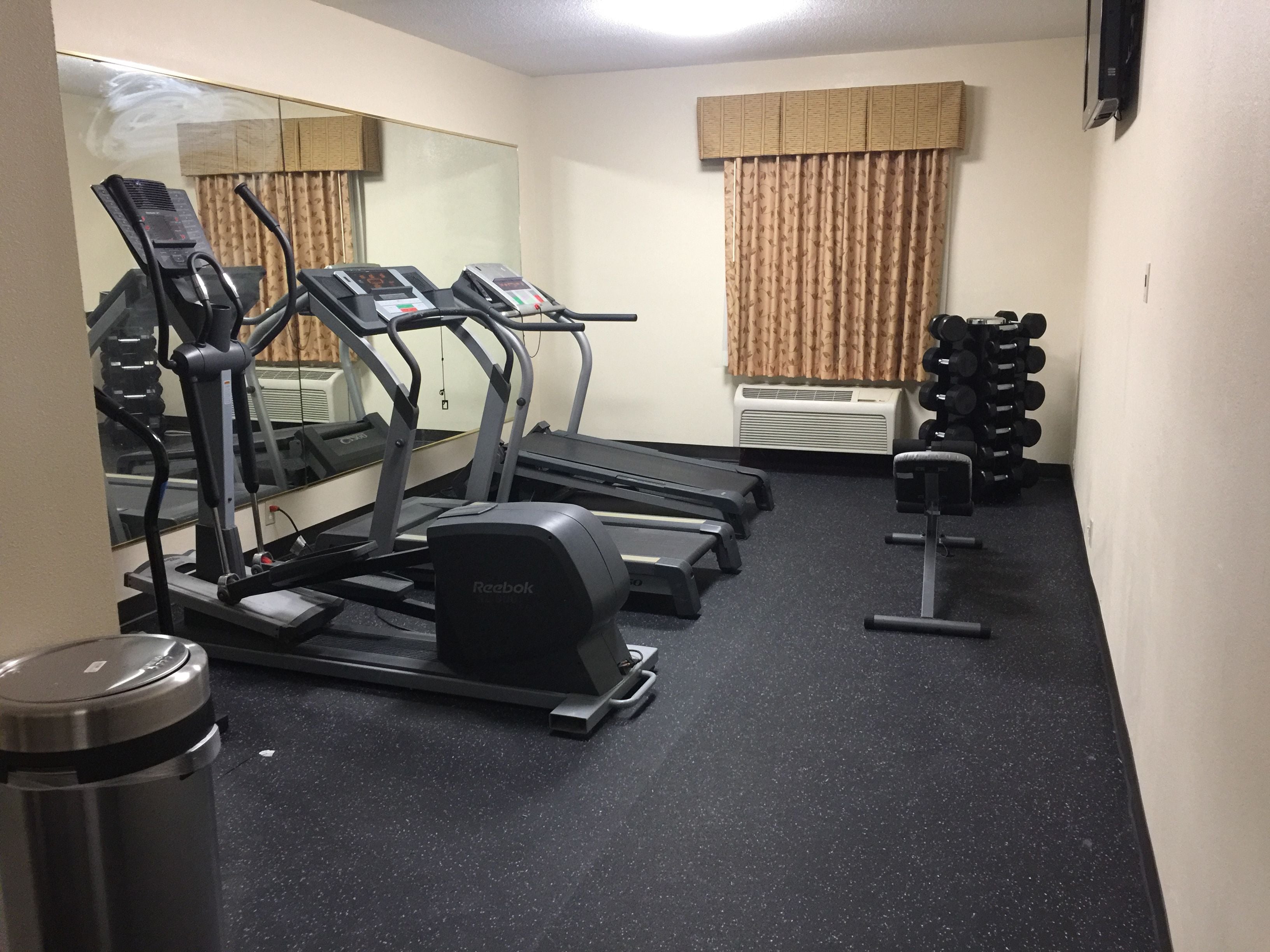 A gym with treadmills, a mirror, a mirror with a reflection, a window with curtains, and a wall-mounted TV.