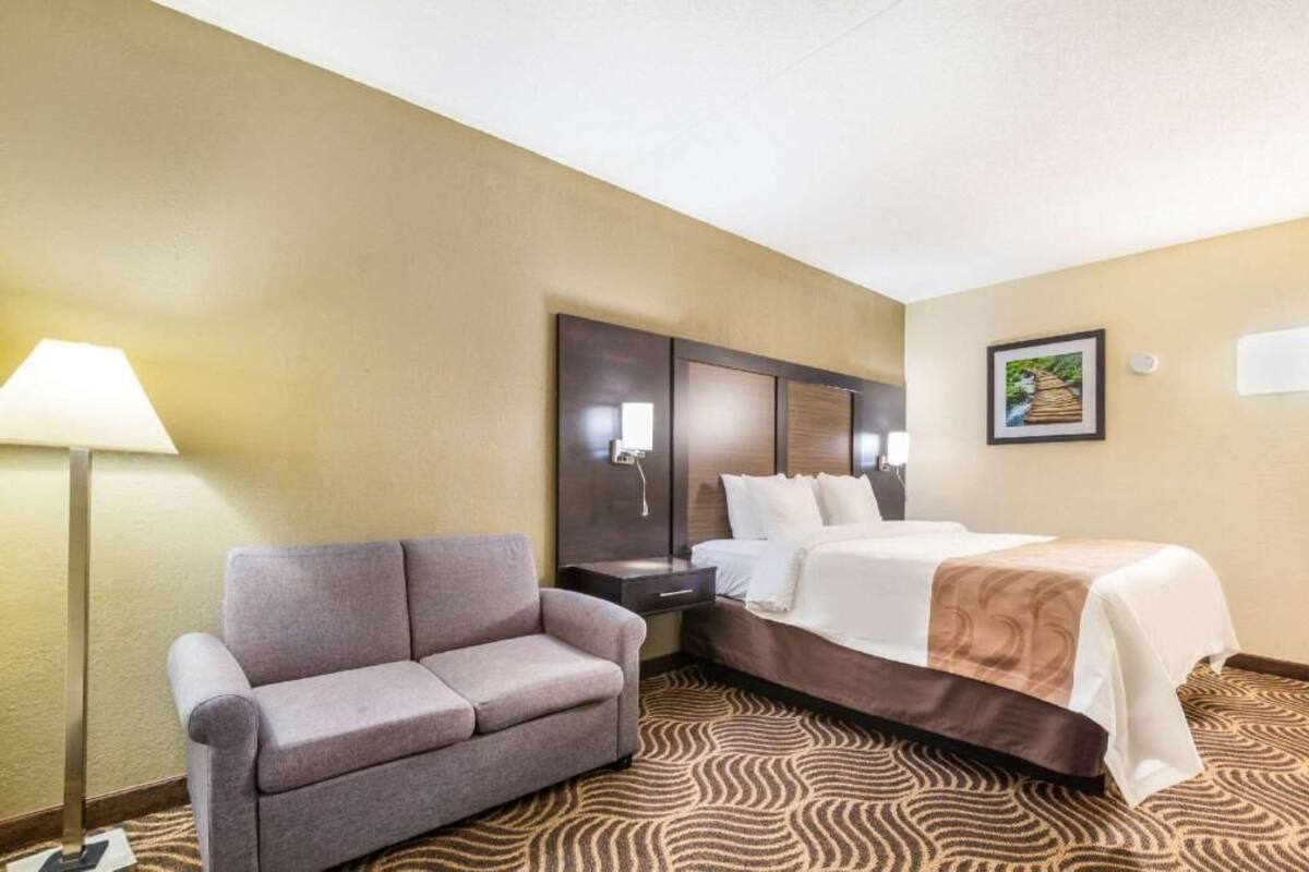 A hotel room with a bed, a sofa, a nightstand, a lamp, and a wall-mounted TV.
