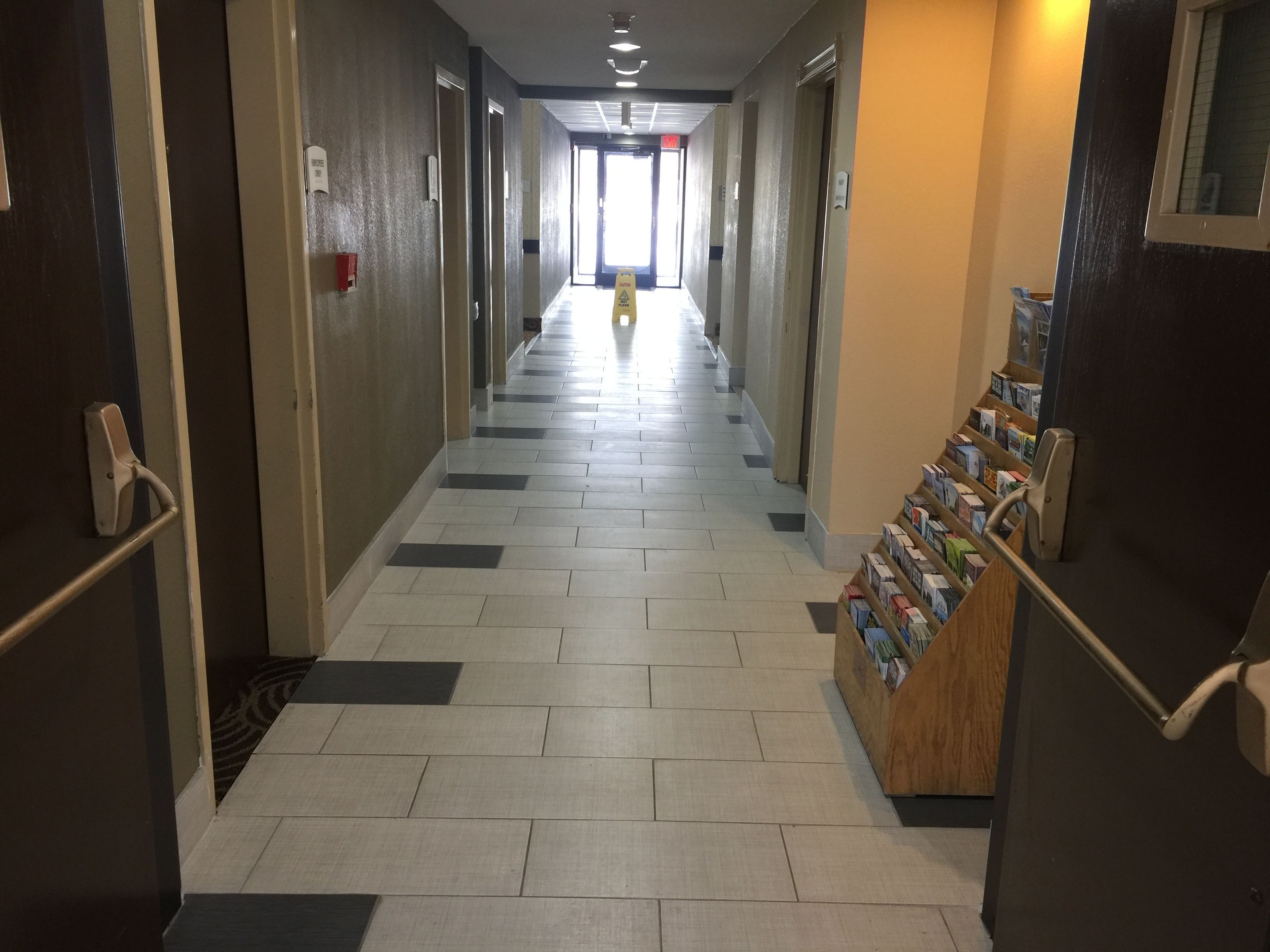 Corredor de hotel com piso de azulejos, portas em ambos os lados e um pequeno quiosque com folhetos à direita.