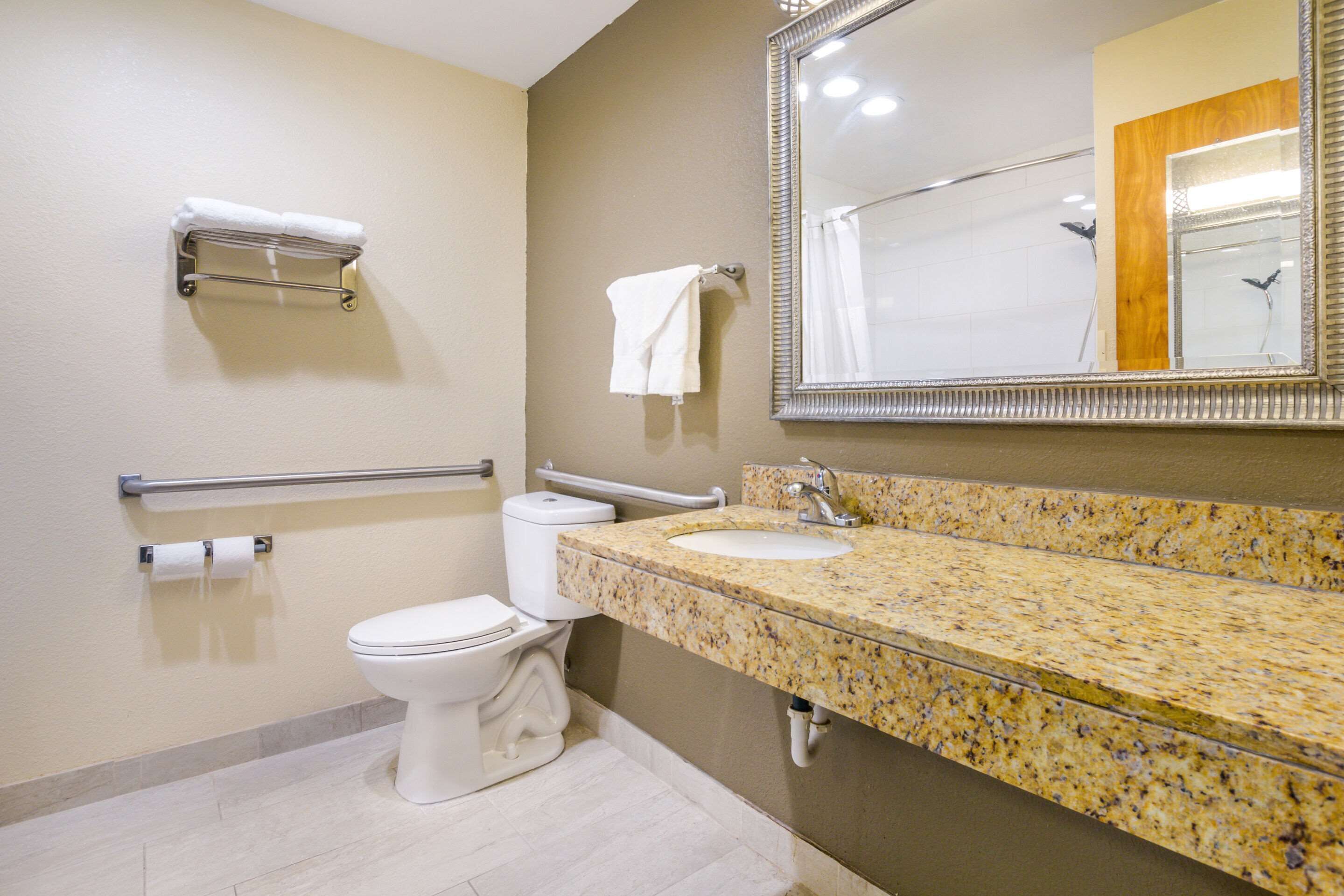 Un baño con inodoro, lavamanos, espejo, toallero y barra de apoyo.