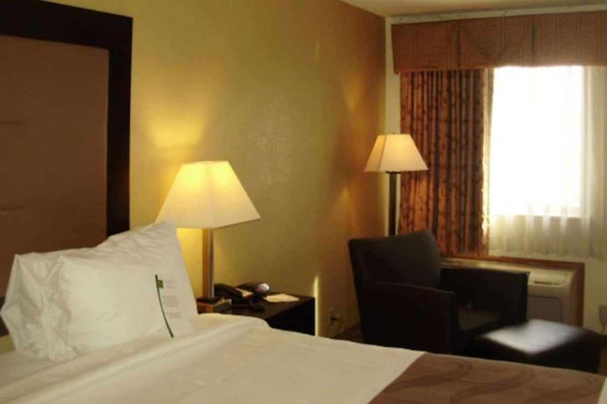A hotel room with a bed, two bedside lamps, a chair, a desk, and a window with curtains.