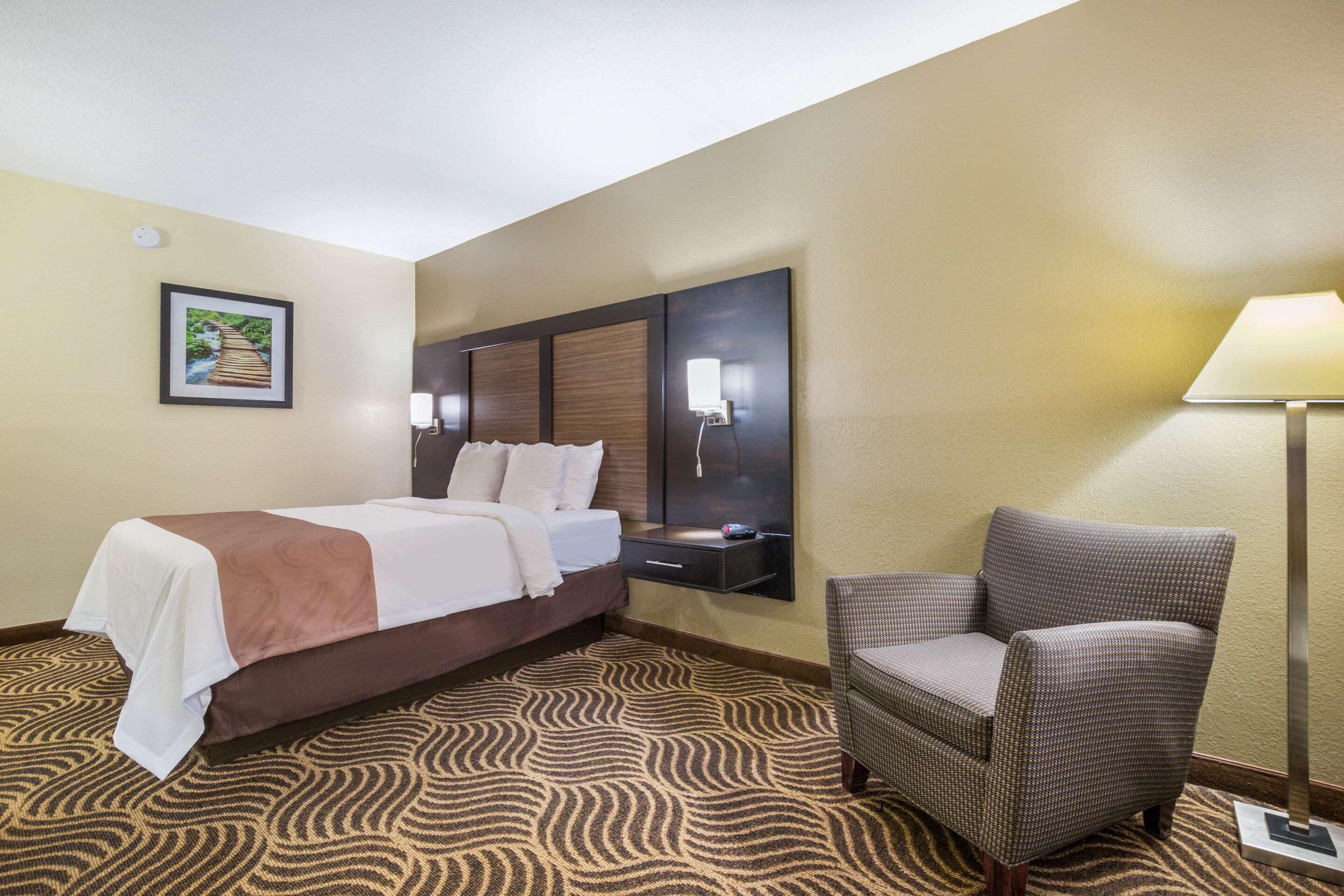 A hotel room with a bed, a chair, a nightstand, and a wall-mounted lamp.