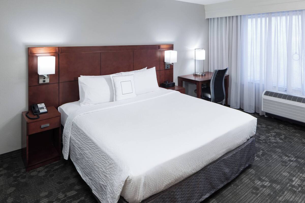 Een hotelkamer met een groot bed, nachtlampjes, een bureau en een raam met gordijnen.
