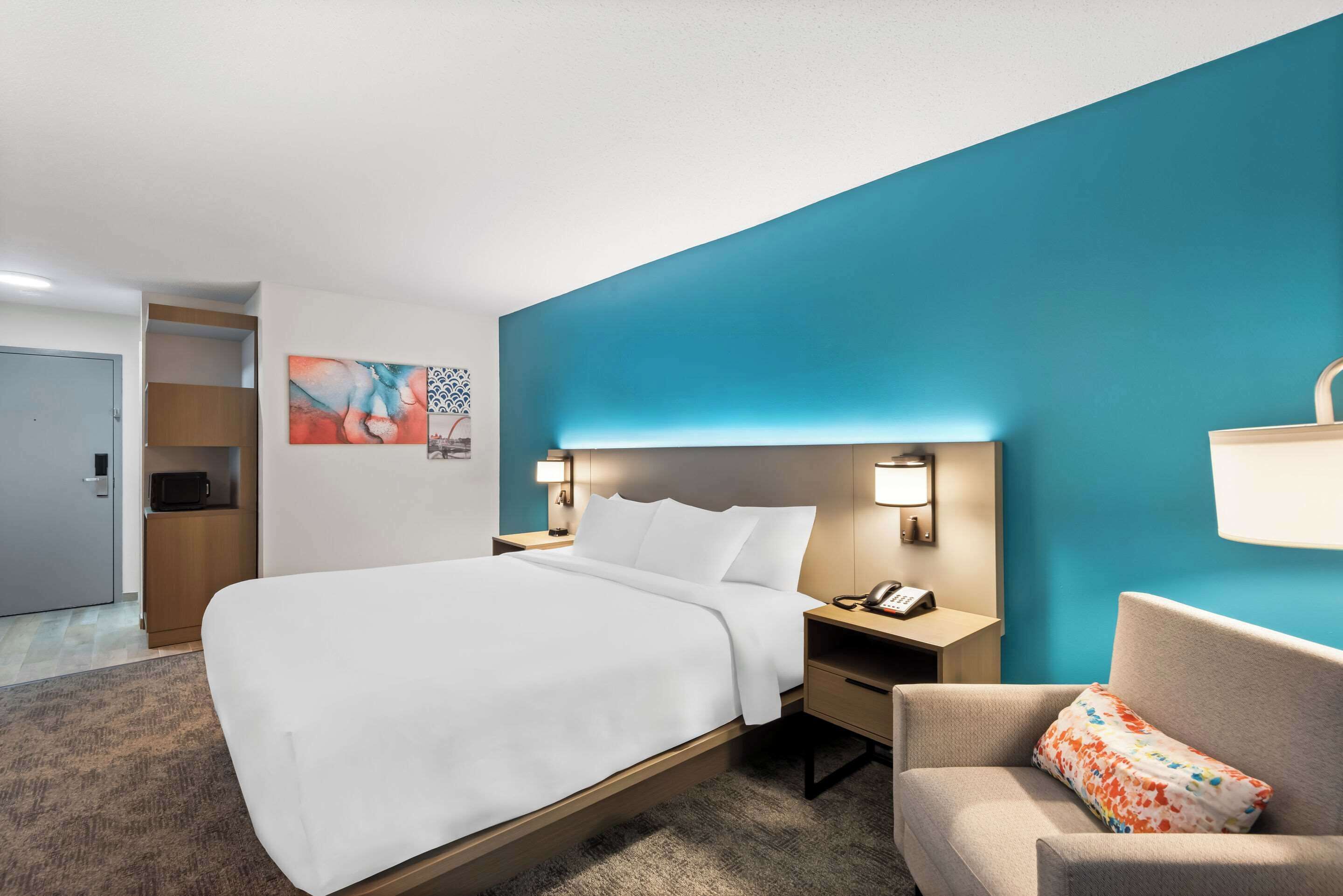 A hotel room with a bed, a nightstand with a lamp, a chair, and a wall with a blue accent.