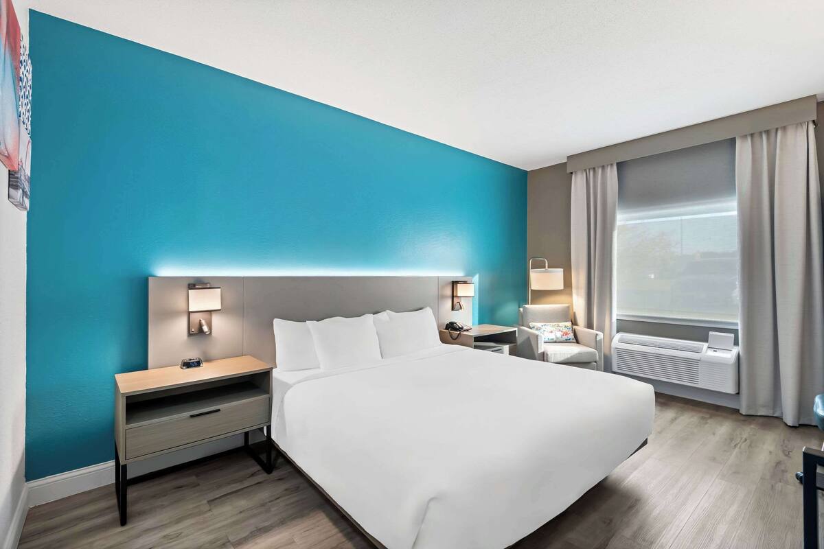 A modern hotel room with a large bed, a bedside table, a nightstand, a desk, a chair, a window with curtains, and a wall-mounted air conditioner.