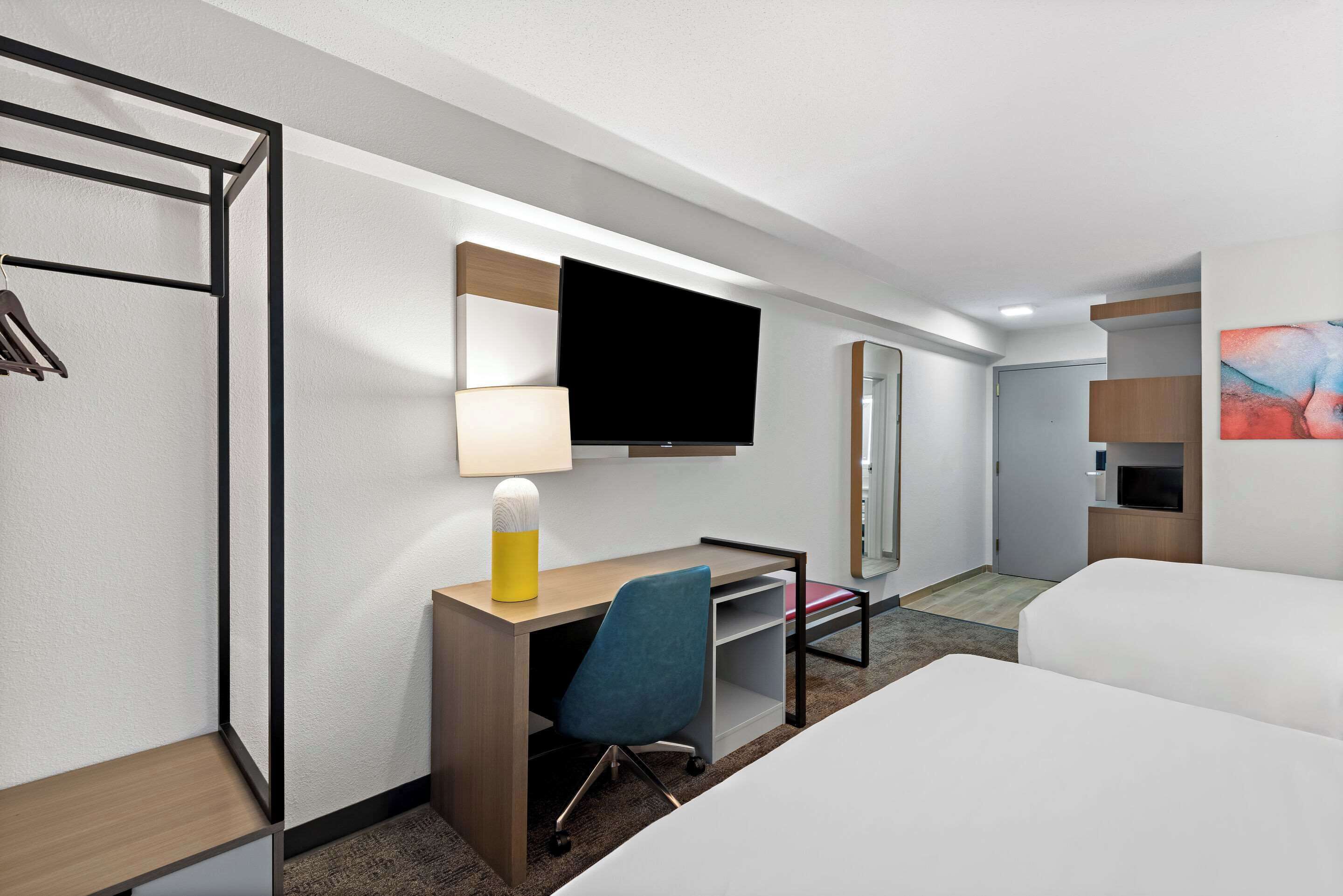 A hotel room with a bed, a desk with a chair, a TV, a lamp, a mirror, and a wardrobe.