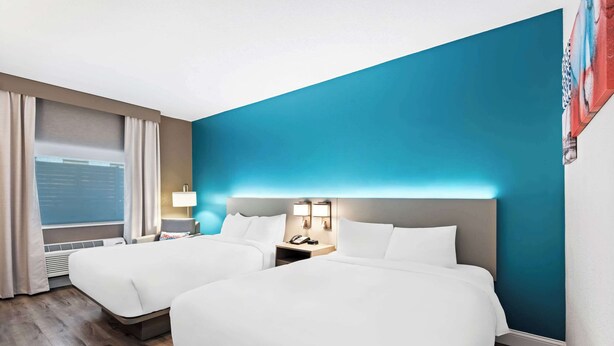 Una camera d'albergo con due letti, un'ampia finestra con tende, un comodino con lampada e una parete decorata in blu.