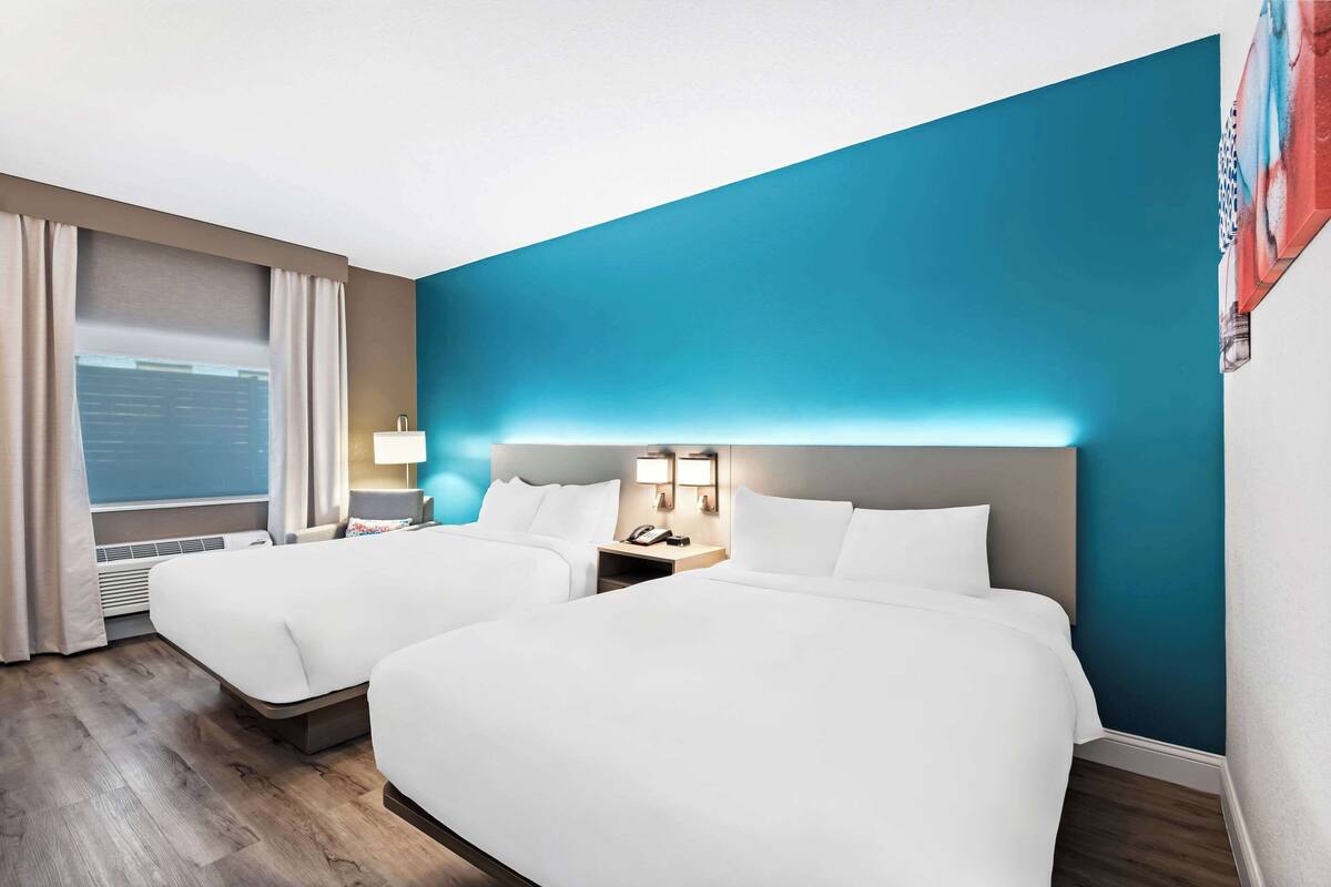 A hotel room with two beds, a large window with curtains, a nightstand with a lamp, and a blue accent wall.