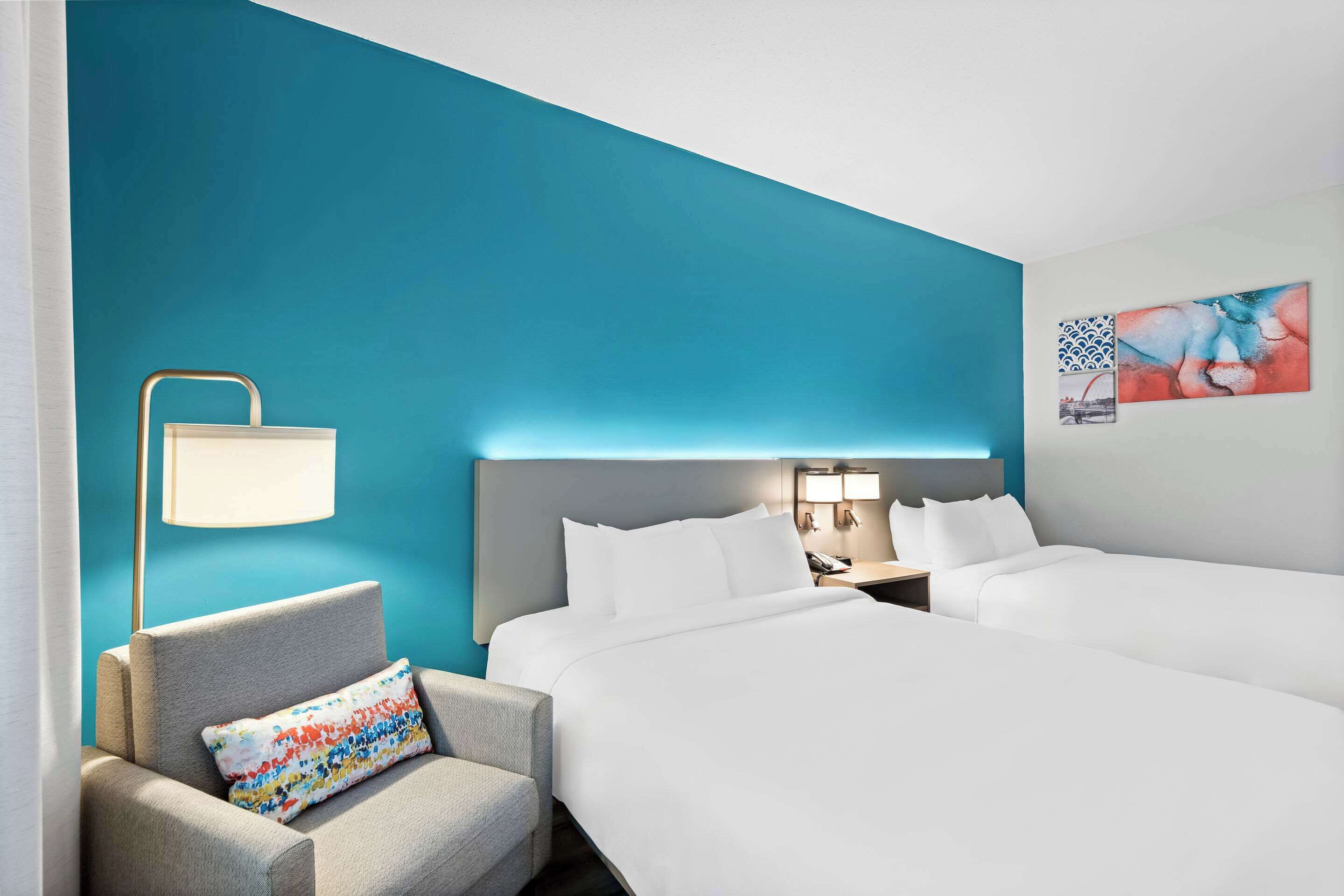Habitación de hotel con dos camas, una silla, una mesita de noche, una lámpara y una obra de arte en la pared.
