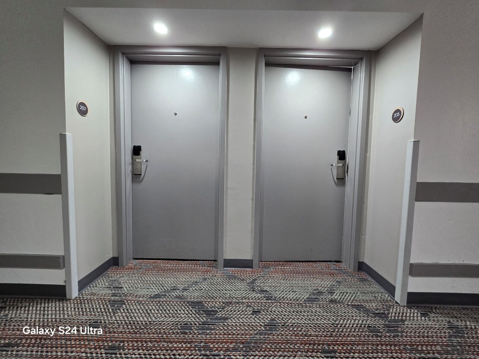 Un pasillo con dos puertas, cada una con un número. El suelo tiene una alfombra estampada.