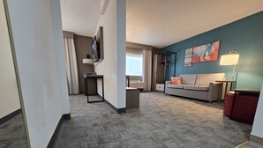 Camera d'albergo con tappeto grigio, un divano, una TV a parete e un quadro sopra il divano.