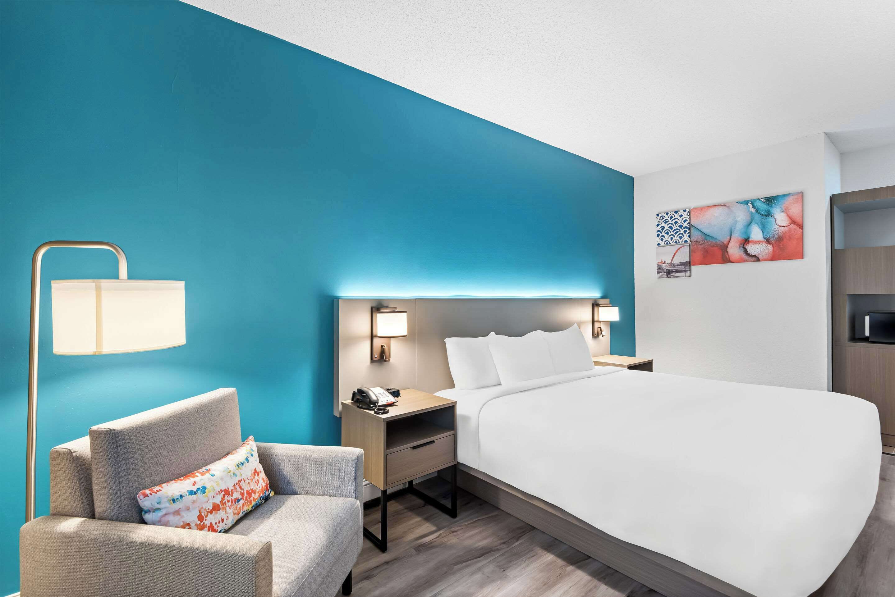A hotel room with a large bed, a chair, a nightstand with a lamp, and a wall with artwork.