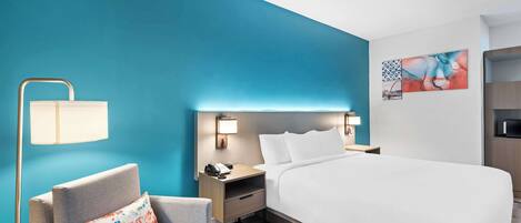 A hotel room with a large bed, a chair, a nightstand with a lamp, and a wall with artwork.