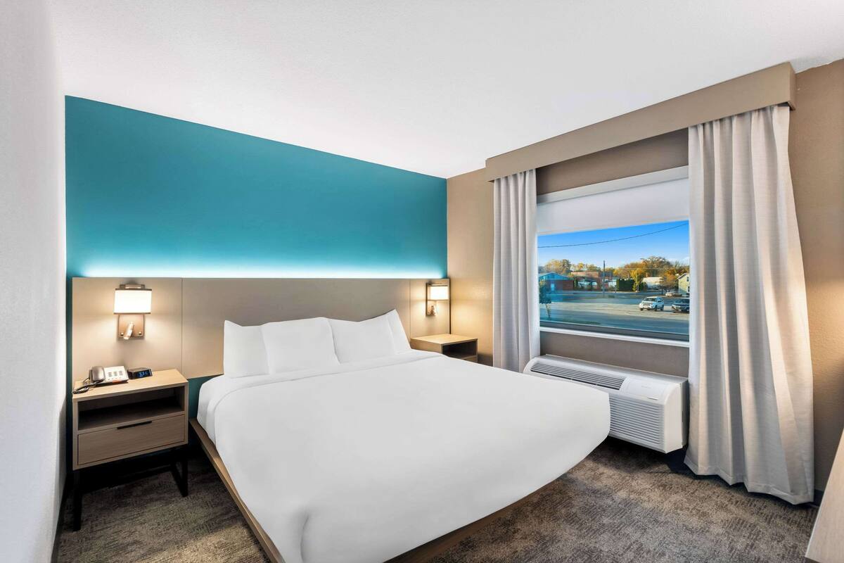 A hotel room with a large bed, a nightstand with a lamp, a window with curtains, and a view of the outside.