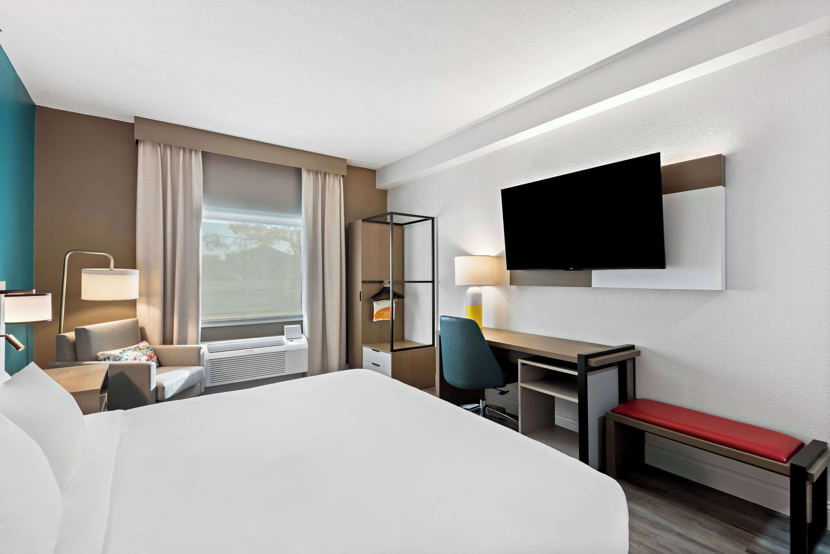 A hotel room with a large bed, a desk with a chair, a television mounted on the wall, and a window with curtains.