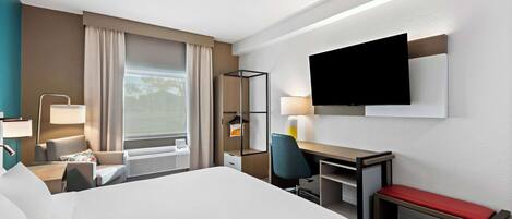 Habitación de hotel con una cama grande, un escritorio con silla, un televisor montado en la pared y una ventana con cortinas.