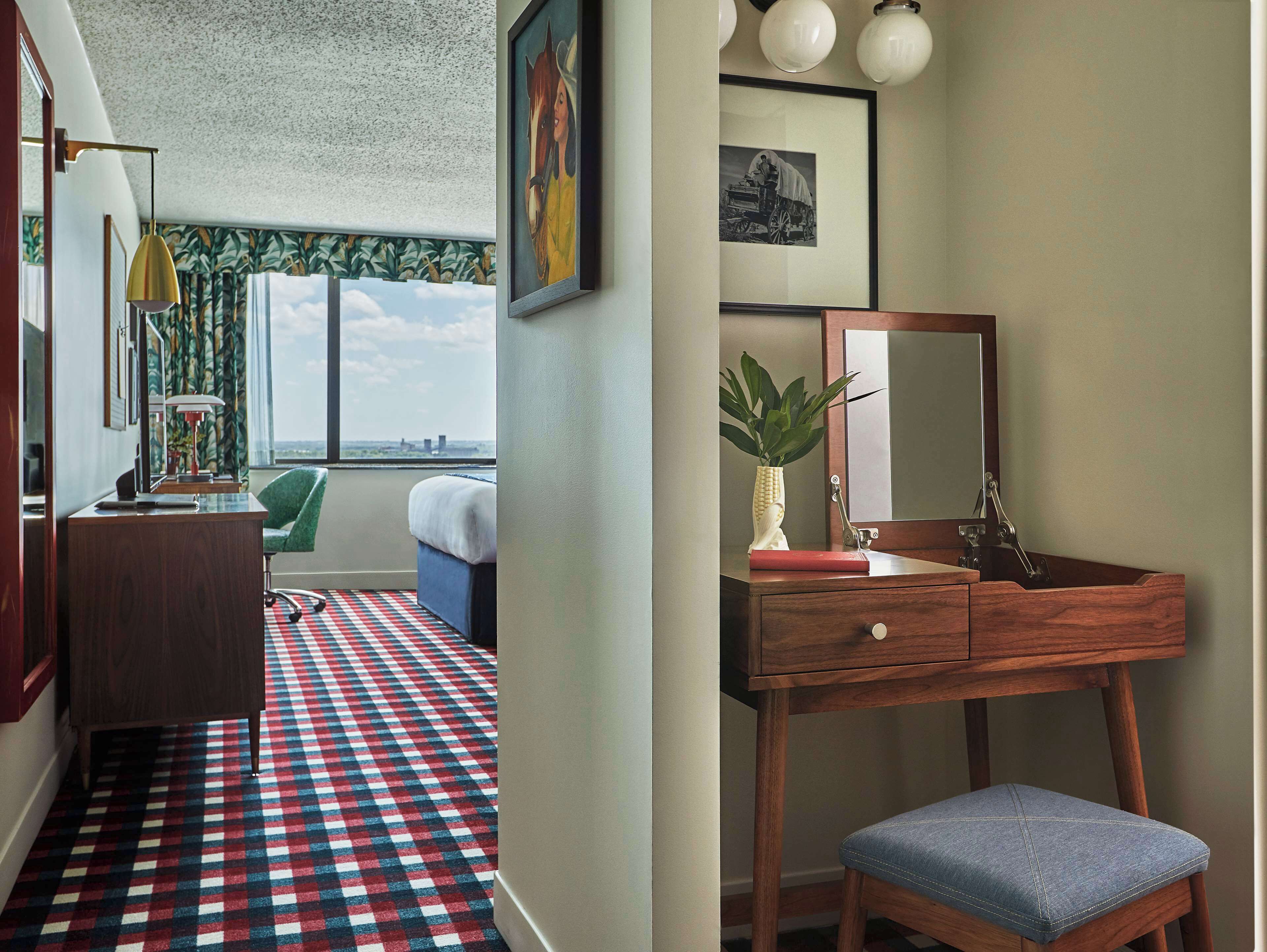A hotel room with a checkered floor, a desk with a lamp, a chair, a bed, a window with a view, a vanity with a mirror, and a plant.