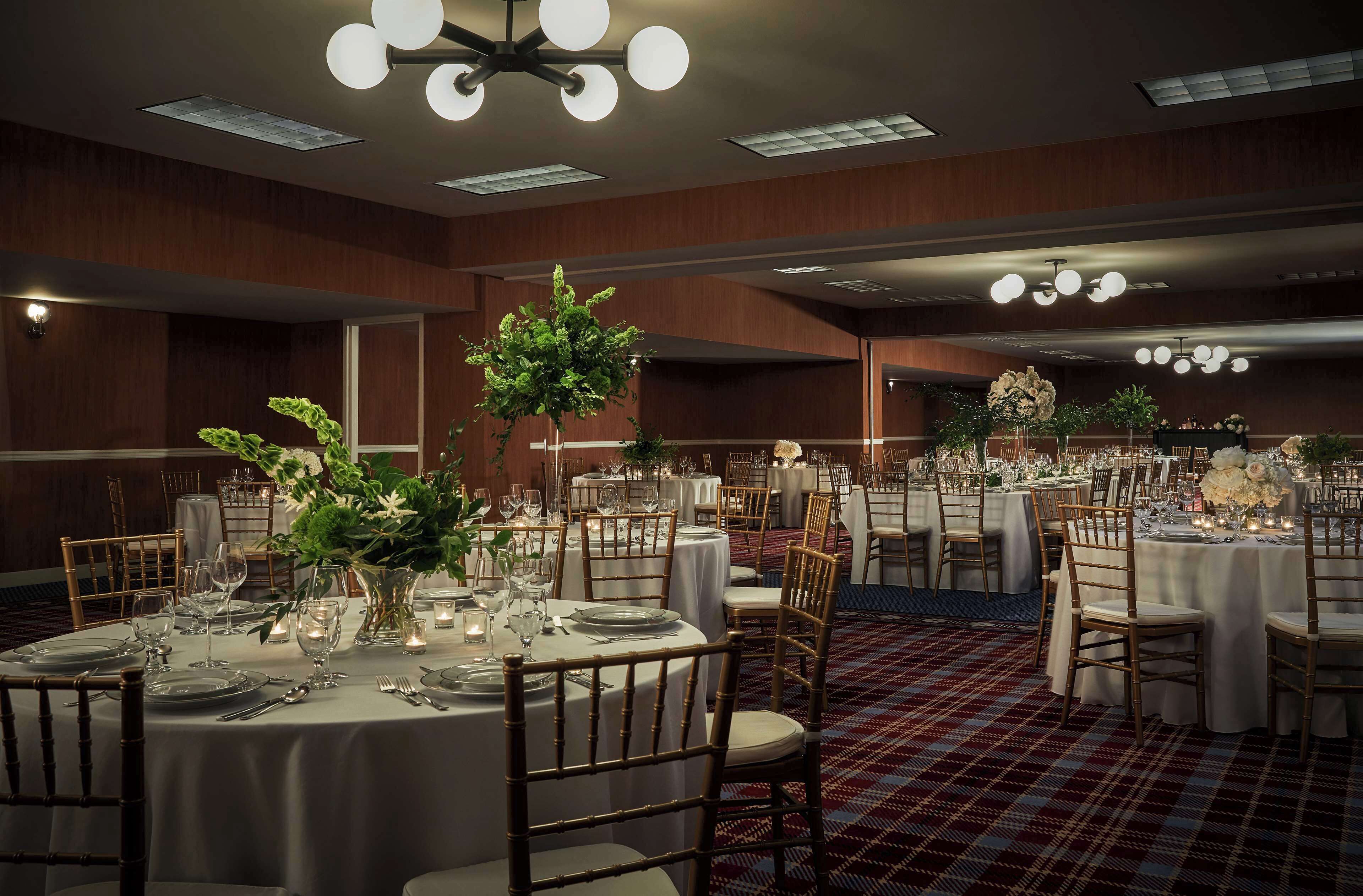 A banquet hall with round tables, white tablecloths, and wooden chairs.