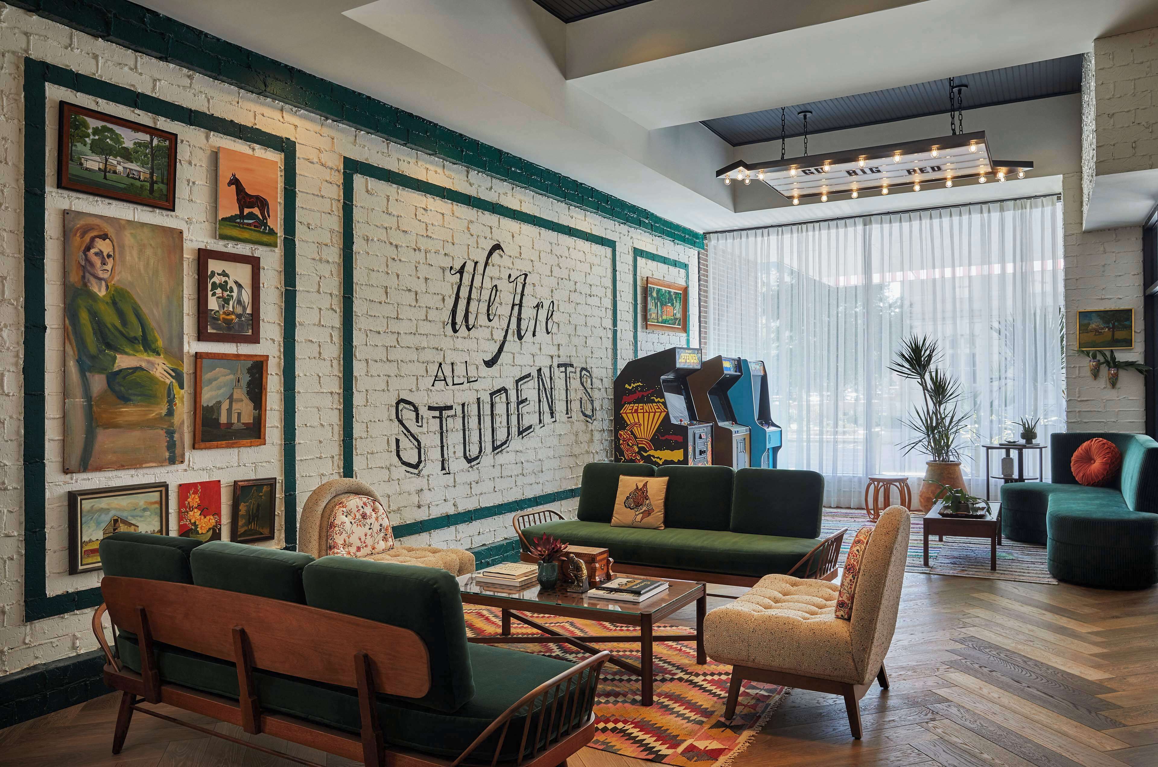 A cozy living room with a brick wall, green sofas, and a large mural reading "We Are All Students."
