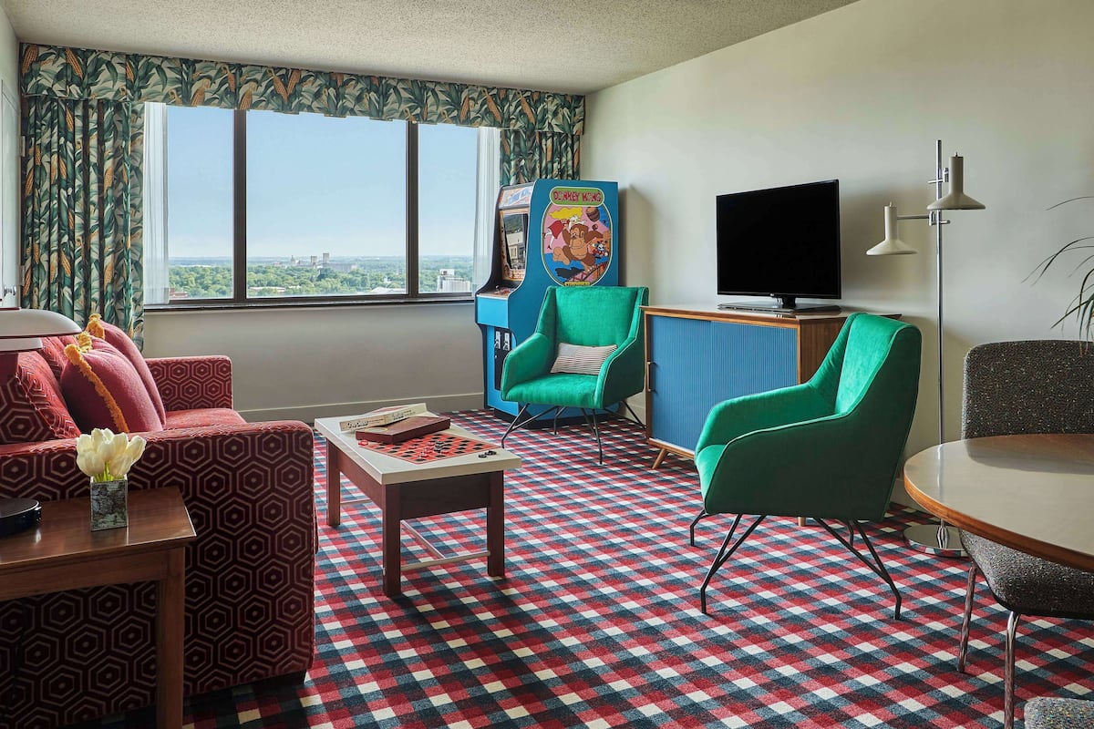 A living room with a checkered carpet, a green armchair, a red patterned sofa, a small table with a board game, a television, and a Nintendo arcade machine.