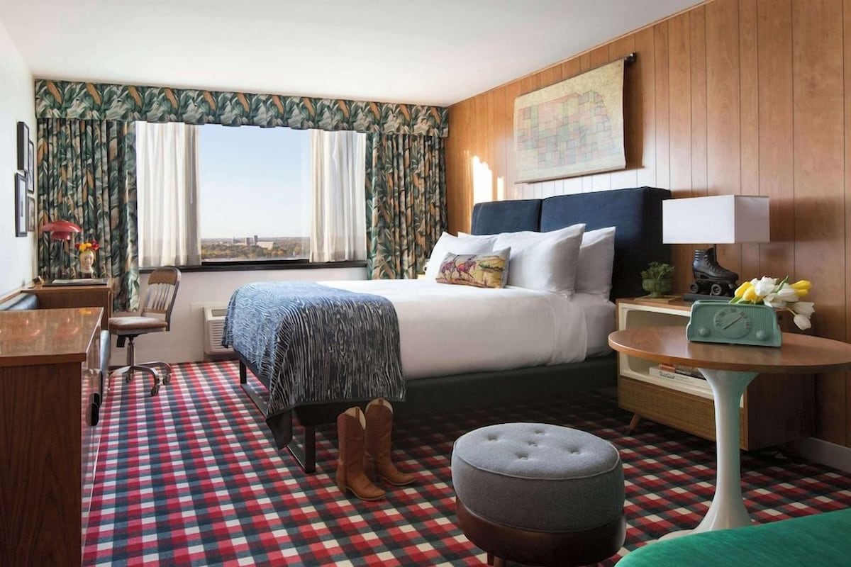 A hotel room with a bed, a desk, a chair, a small table, and a window with curtains.