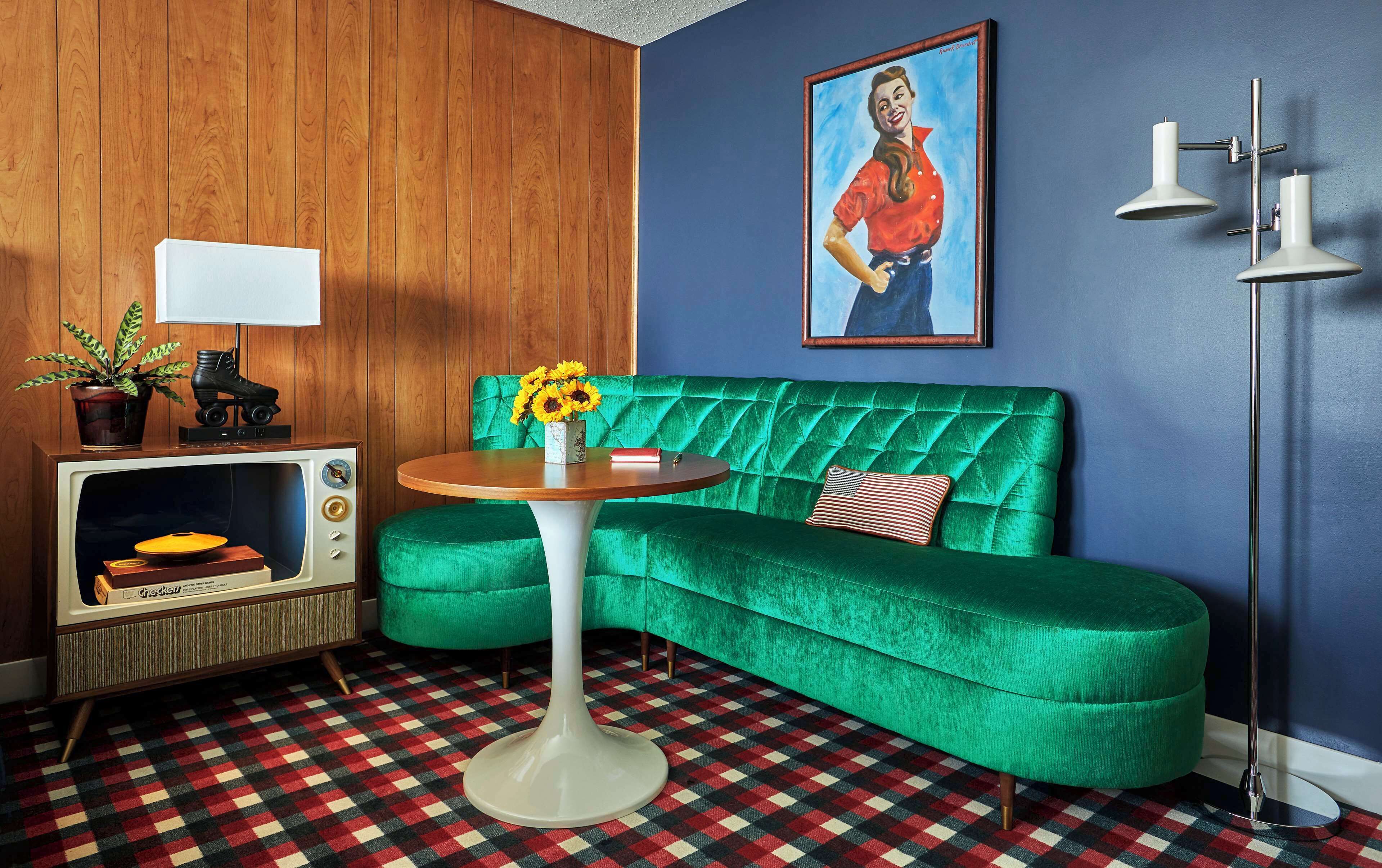Una habitación con un sofá de terciopelo verde, un televisor antiguo, una alfombra estampada y un cuadro en la pared.