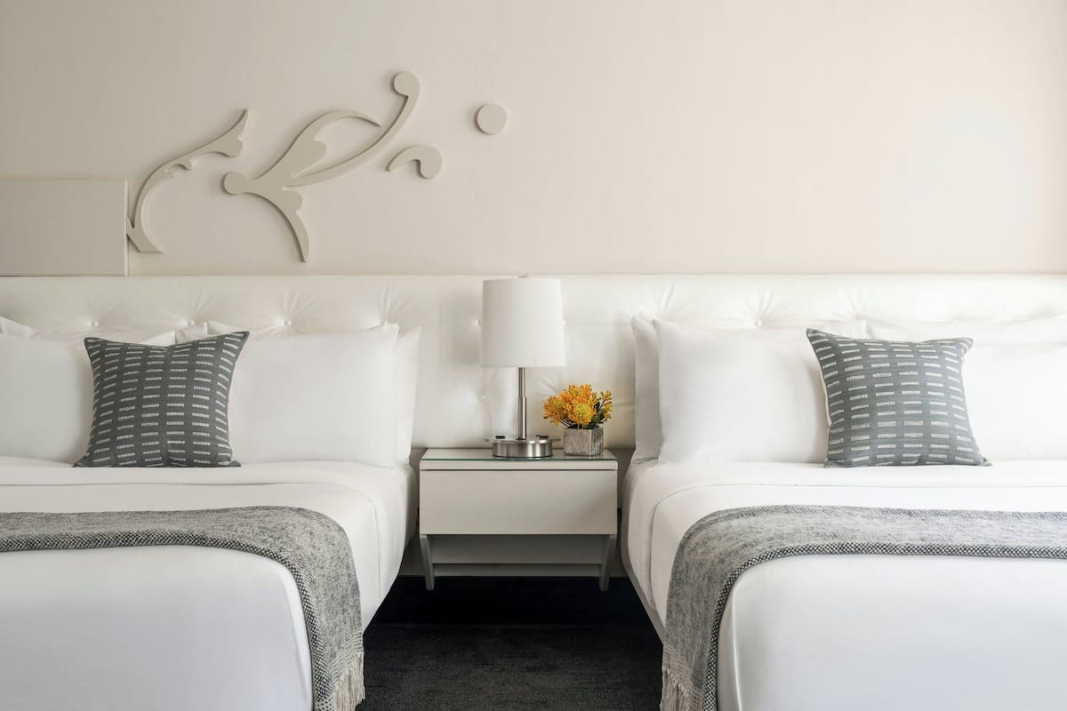 A hotel room with two beds, a nightstand with a lamp and flowers, and a decorative wall piece.