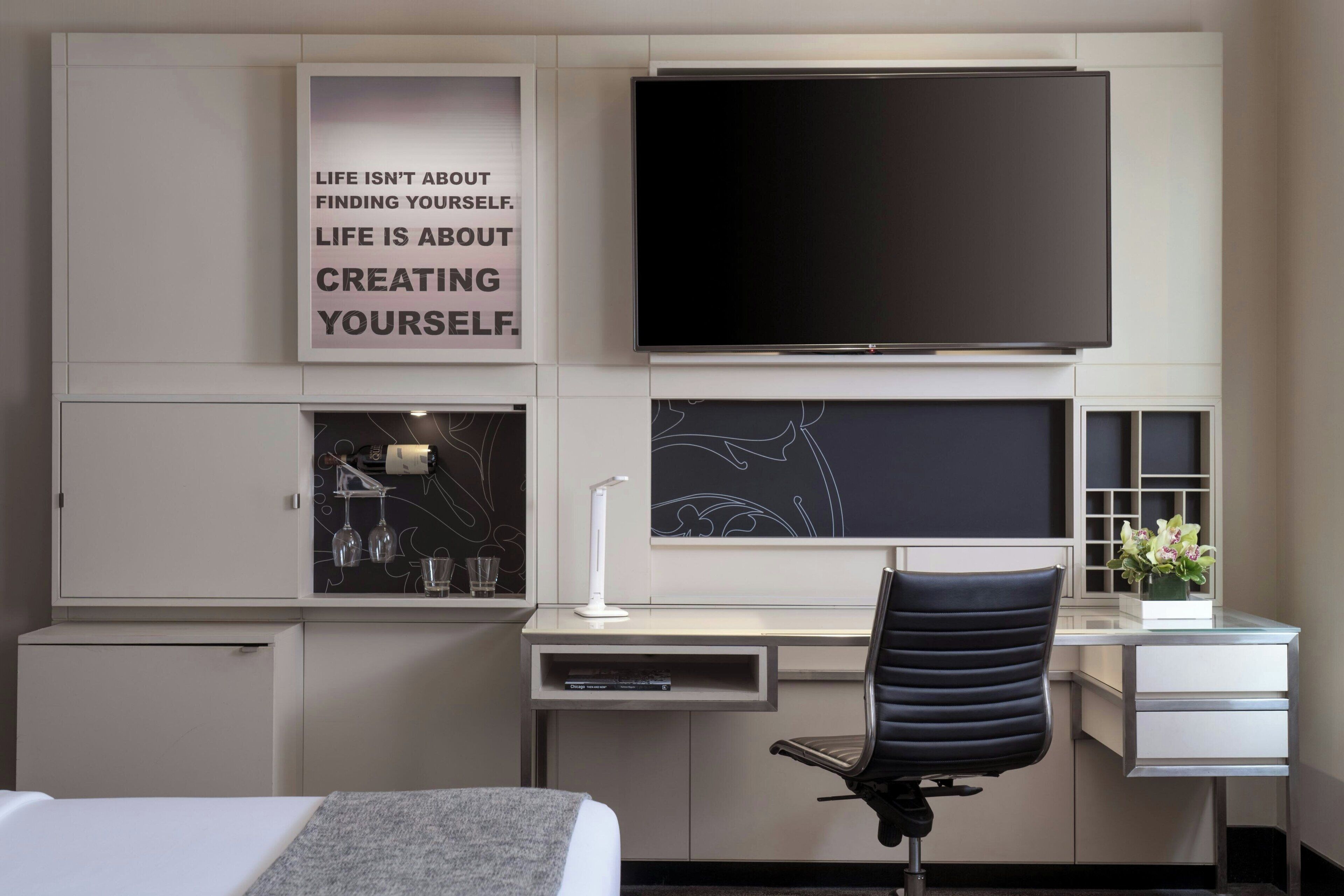 A hotel room with a bed, a desk with a chair, a TV, a wall with motivational quote, and a small cabinet with decorative items.