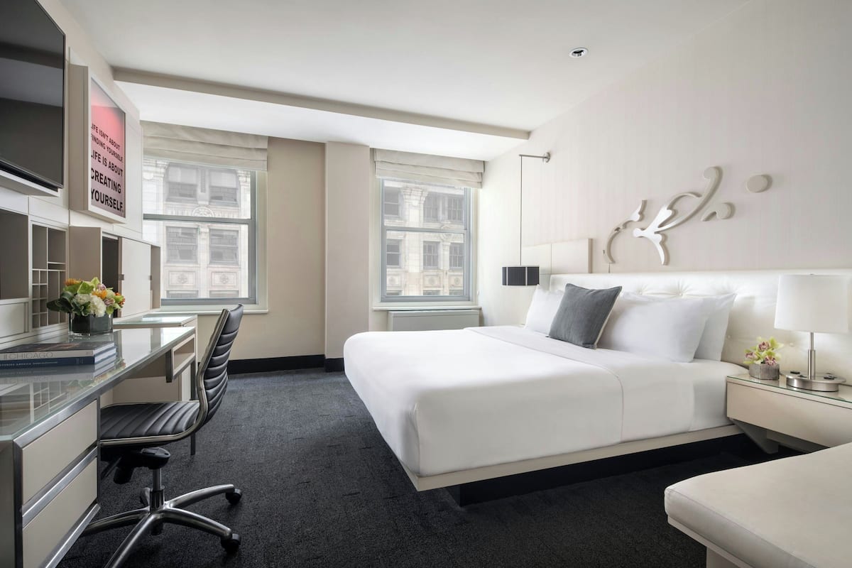 A hotel room with a bed, desk, chair, and a view of the city through large windows.