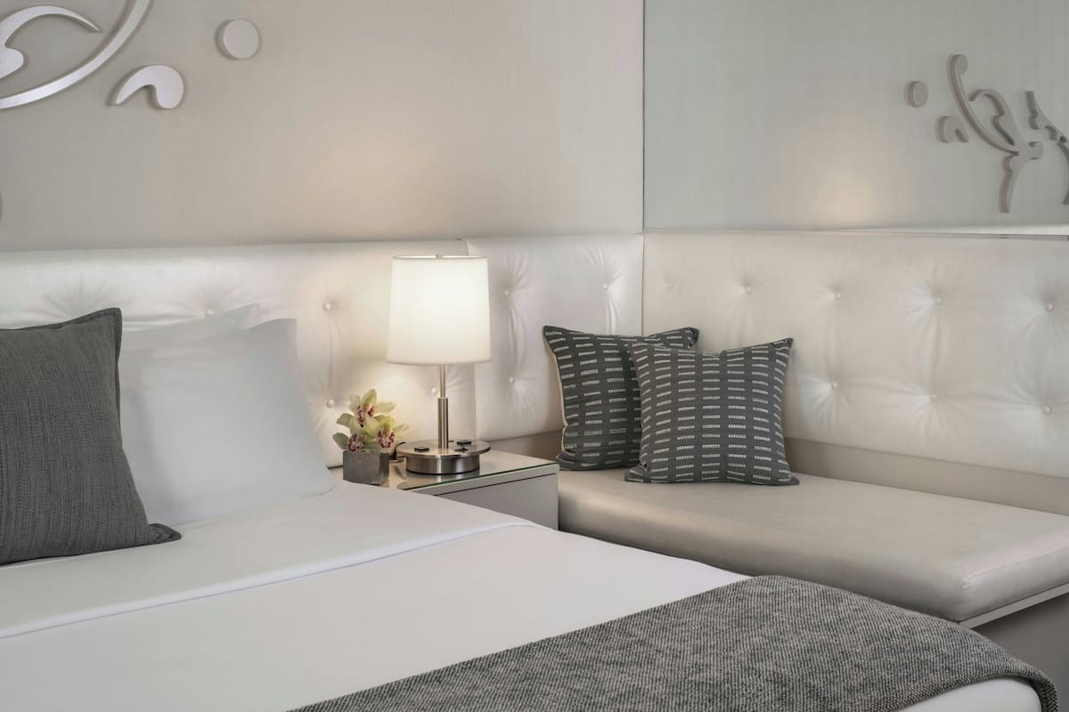 A modern hotel room with a white bed, grey and patterned pillows, a bedside table with a lamp, and decorative wall art.