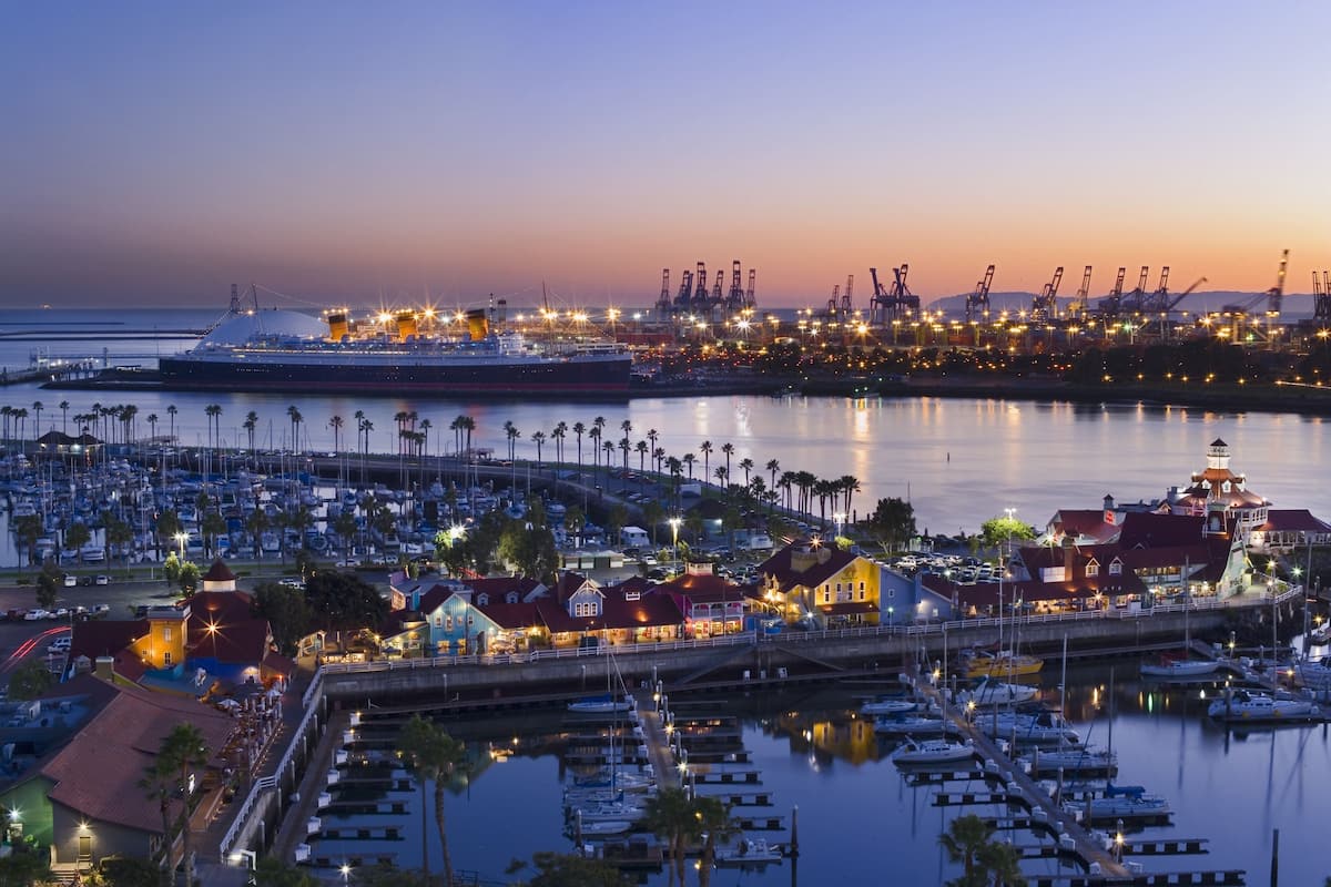 A marina with boats docked, a large ship, and industrial cranes in the background.