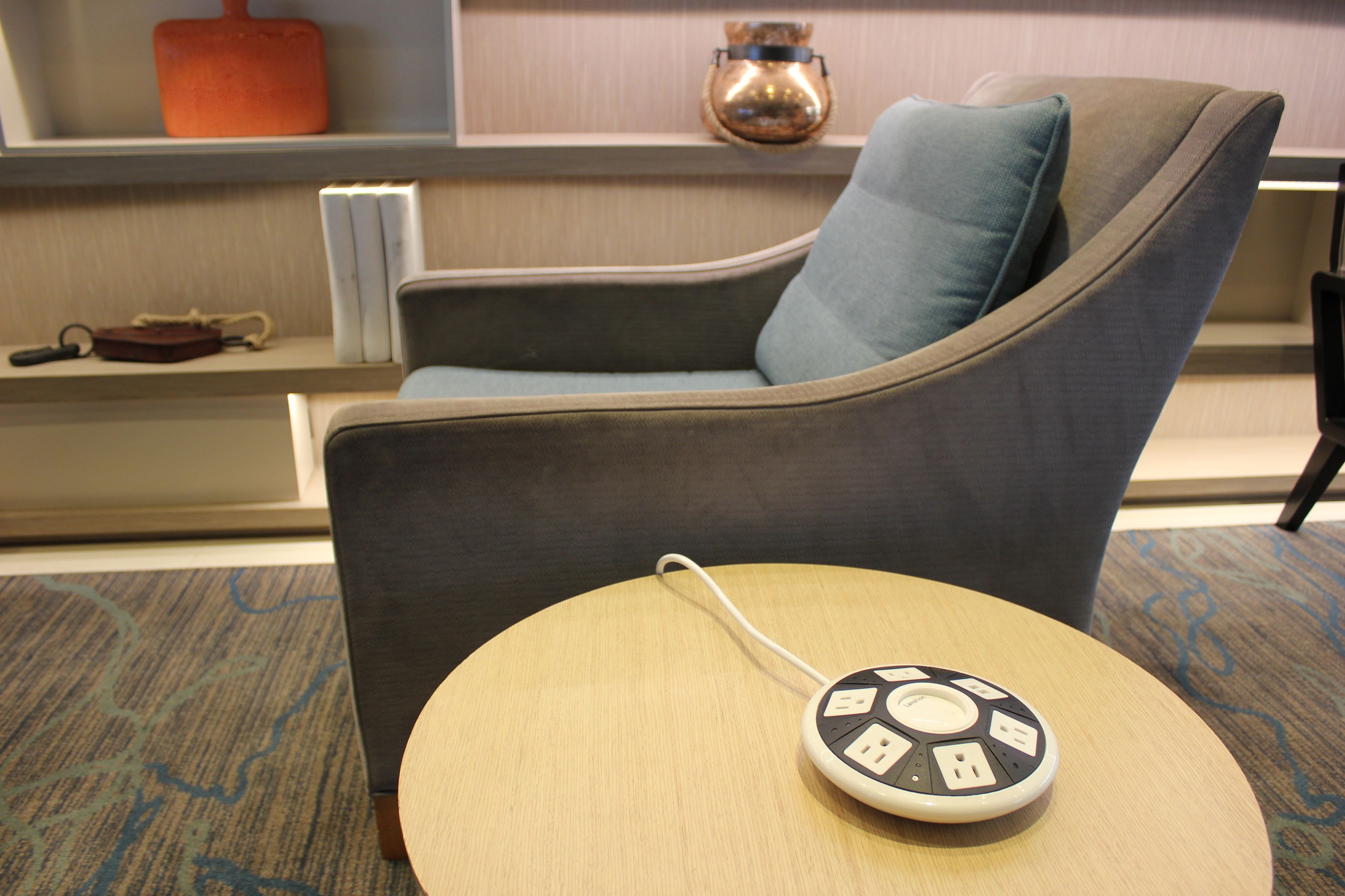 Una habitación de hotel moderna con un sillón gris, una mesa redonda de madera con una regleta eléctrica y una estantería con libros y objetos decorativos.