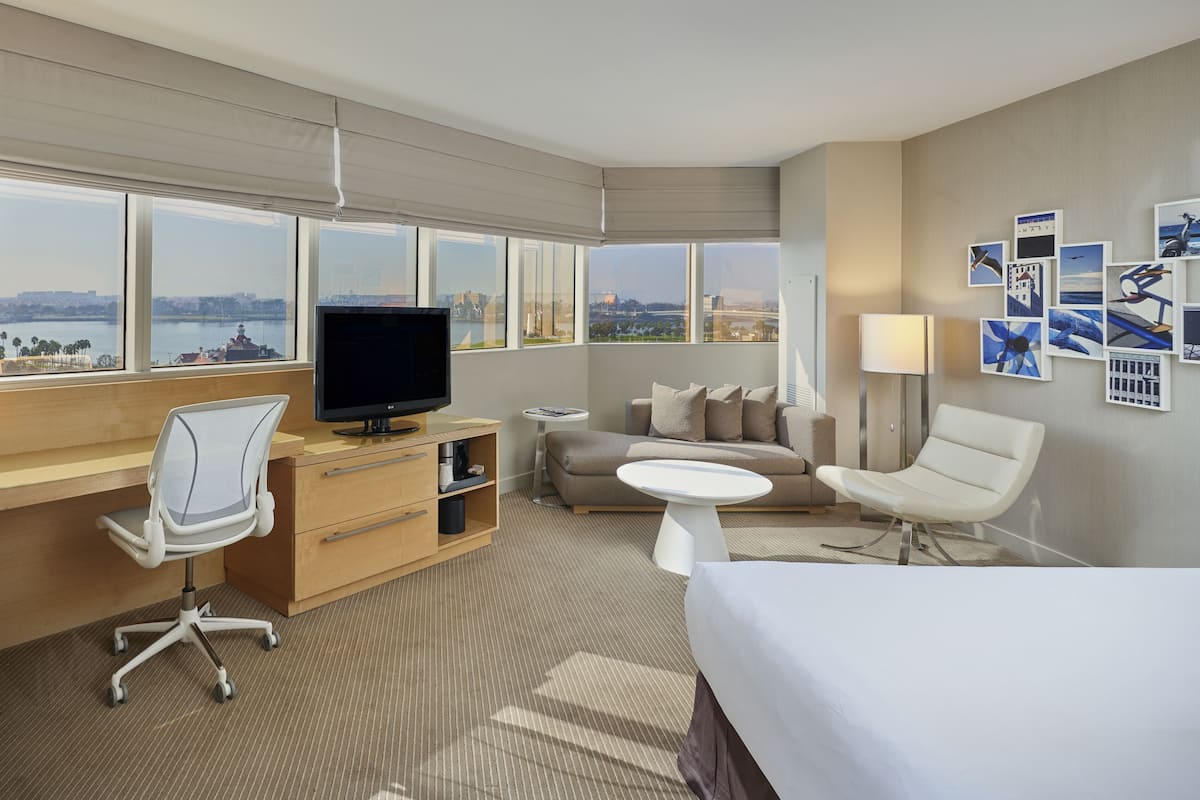 A hotel room with a large window, a desk with a computer, a sofa, a bed, and a small table.