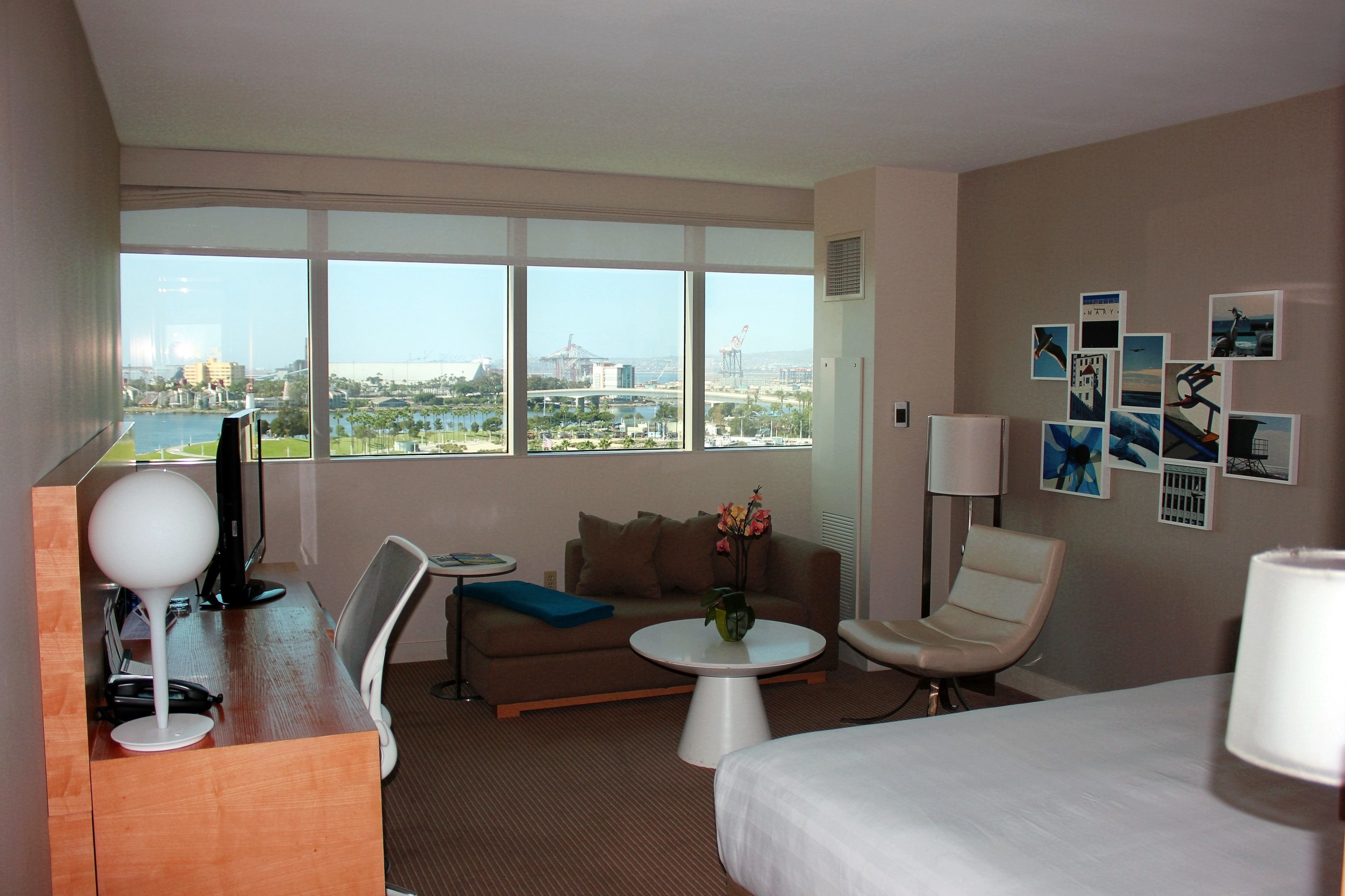 A hotel room with a large window, a sofa, a small table, a desk with a lamp, a chair, and a bed.
