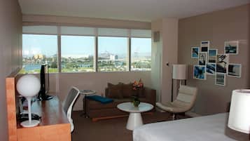 A hotel room with a large window, a sofa, a small table, a desk with a lamp, a chair, and a bed.