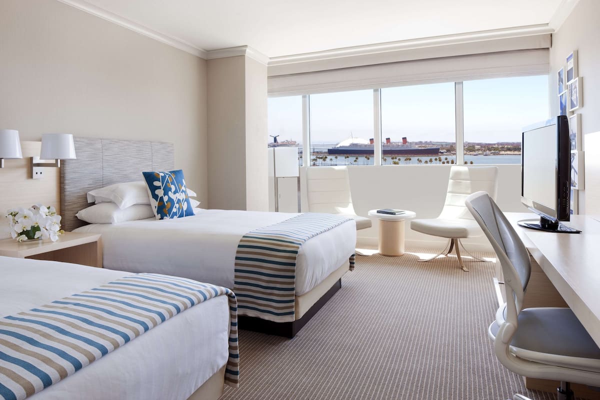 A hotel room with two beds, a desk, a chair, and a large window offering a view of the sea and cityscape.