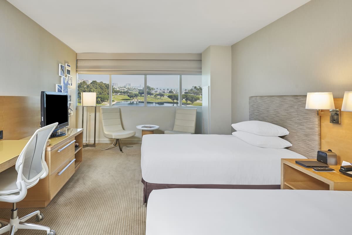 A hotel room with two beds, a desk with a computer, a chair, a lamp, and a view of a park and buildings.