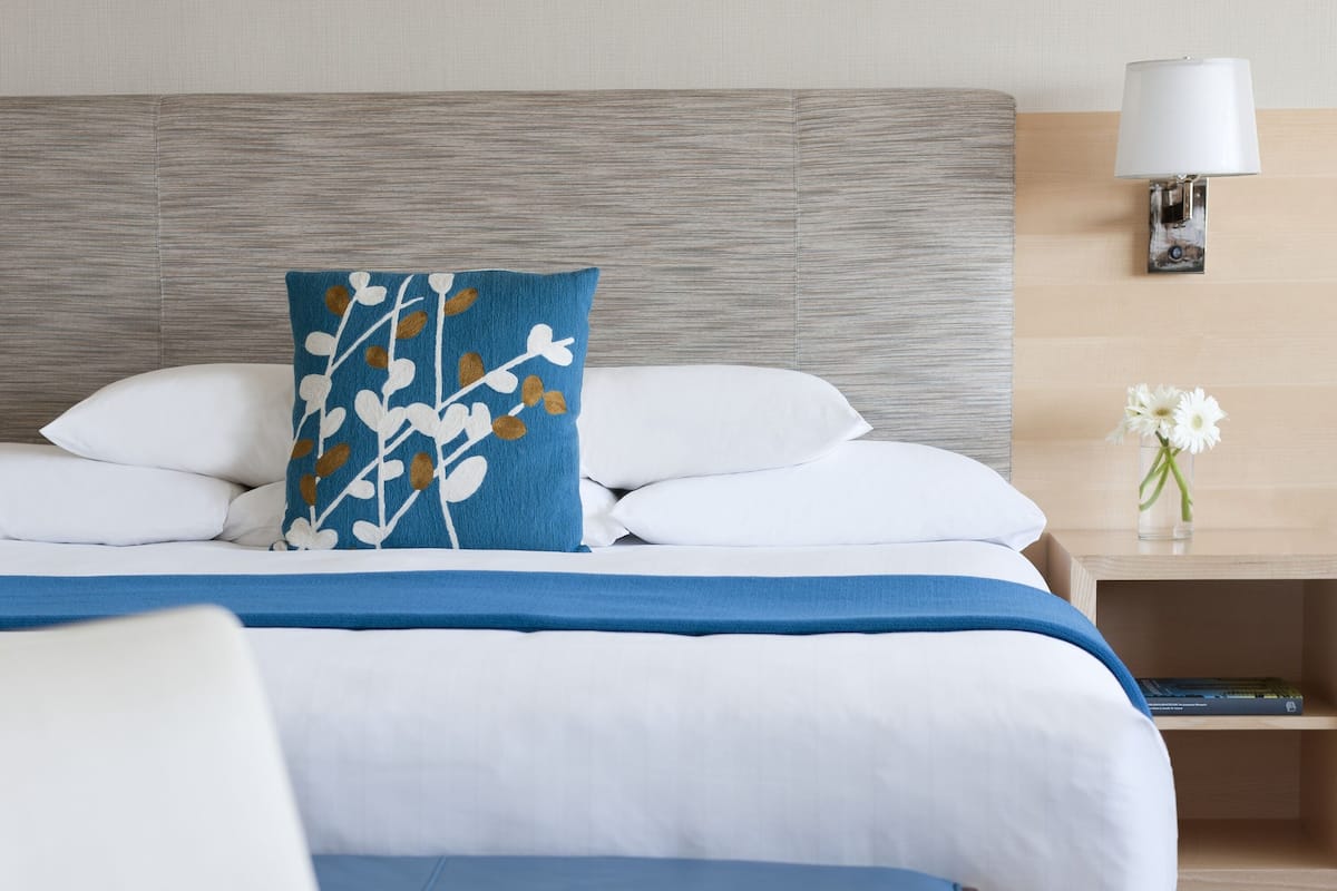 A neatly made bed with white and blue bedding, a decorative pillow, a bedside table with a lamp, and a vase with flowers.
