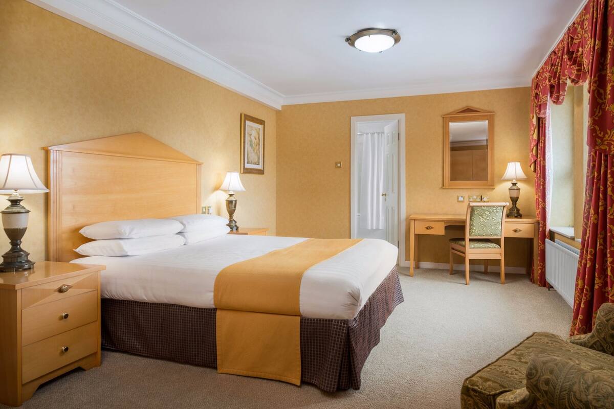 Une chambre d’hôtel comprenant un lit, des tables de chevet, un bureau, une chaise, un miroir et une fenêtre avec des rideaux.