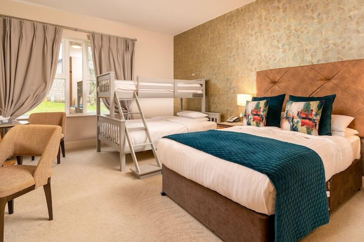Ein Hotelzimmer mit Etagenbett, Schreibtisch mit Stuhl, Nachttisch mit Lampe und einem Fenster mit Vorhängen.