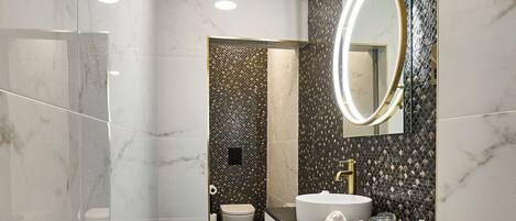 Un baño moderno con tocador de mármol, un espejo redondo y un inodoro visible al fondo.
